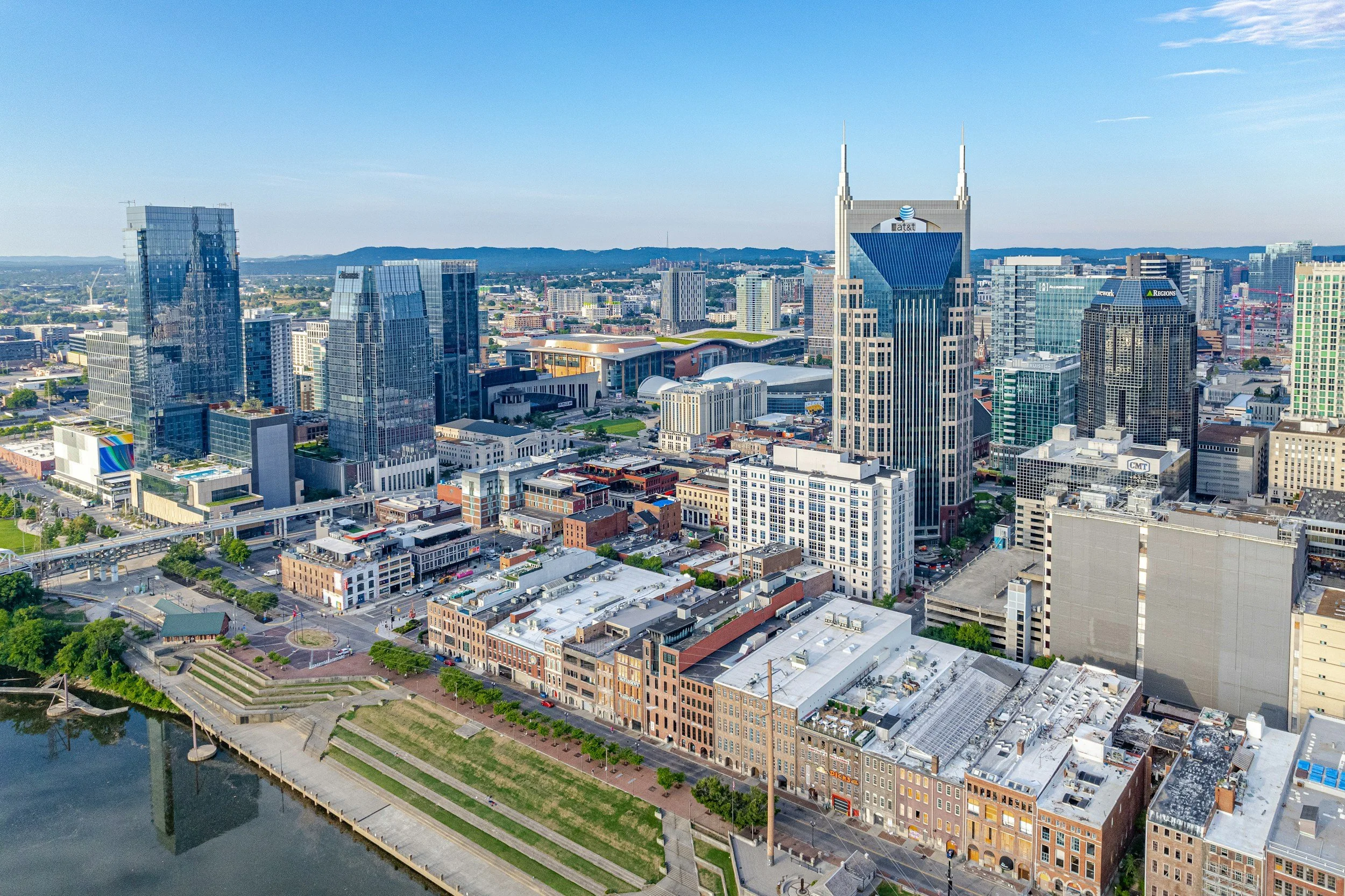 A cityscape with modern skyscrapers along a river, blue sky, and distant hills.