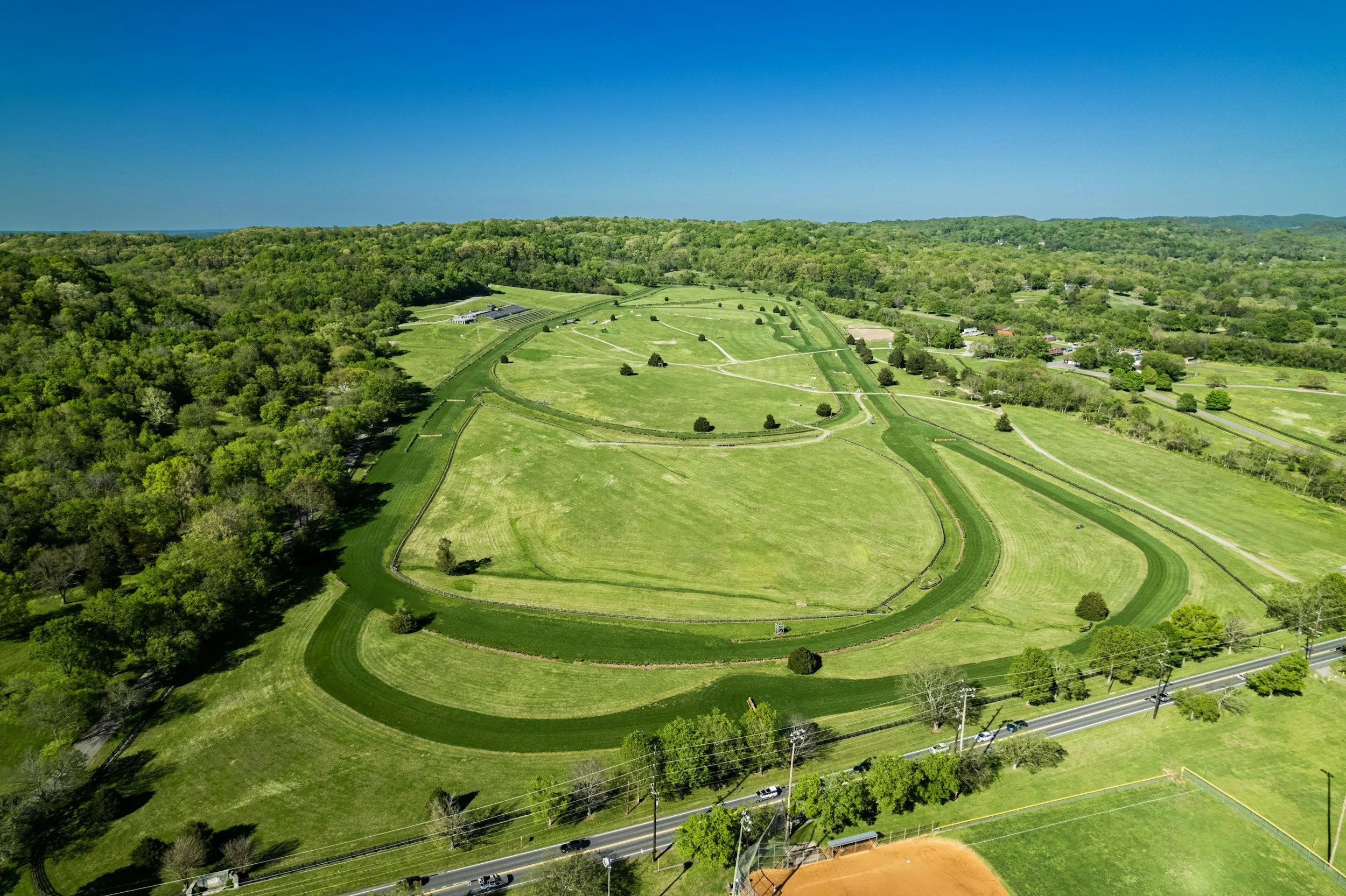 Aerial view of a lush, green landscape with a large open park featuring winding walking trails, trees, and grass. Surrounding the park are densely wooded areas, a few buildings, and a road with vehicles at the bottom.