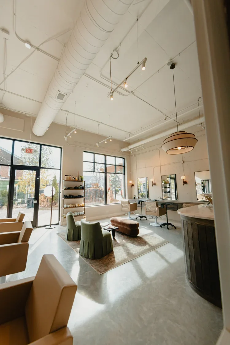Interior of a modern salon with large windows, green and beige chairs, mirrors, and a reception desk, illuminated by hanging light fixtures.
