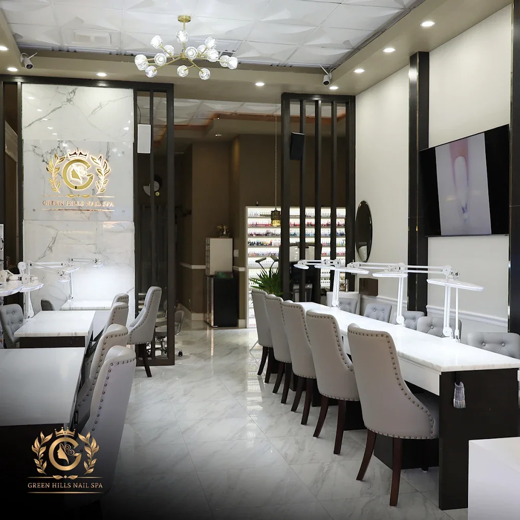 Interior of Green Hills Nail Spa featuring white marble floors, grey upholstered chairs, manicure stations with lamps, a decorative chandelier, a mounted TV, and a wall display of nail polish bottles.