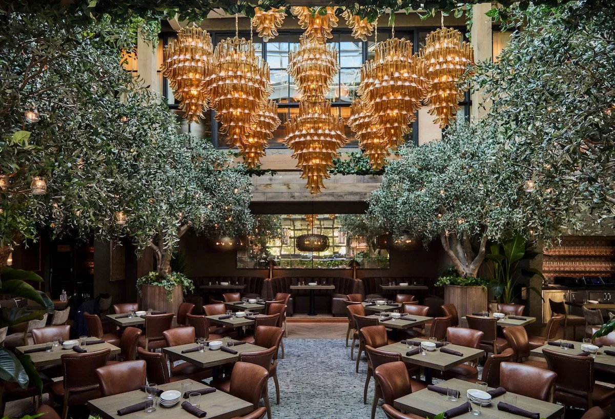 Elegant restaurant interior with a high ceiling, large hanging chandelier, and lush greenery including trees and plants, arranged around tables set for dining.