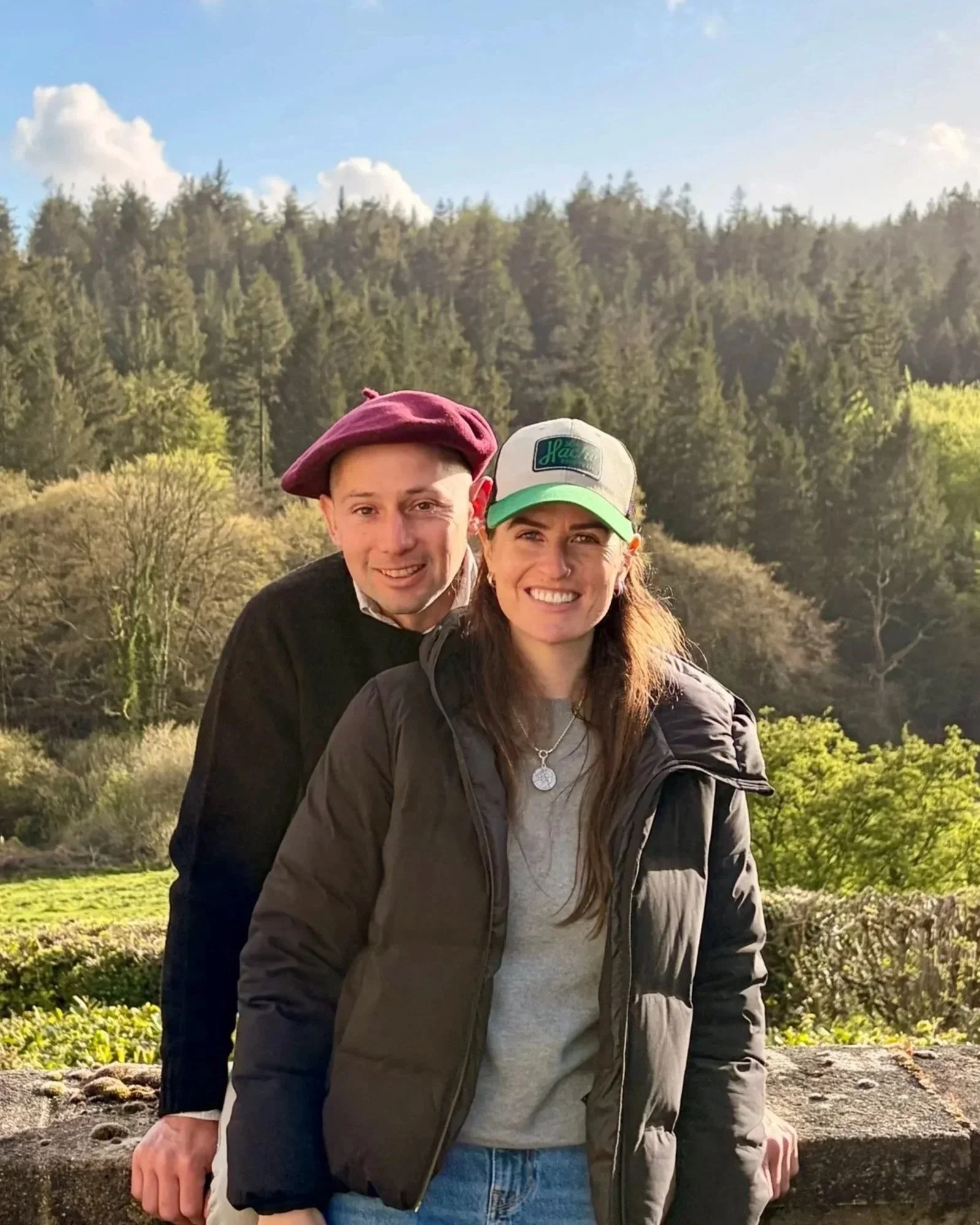Two smiling people outdoors with trees and a blue sky in the background.