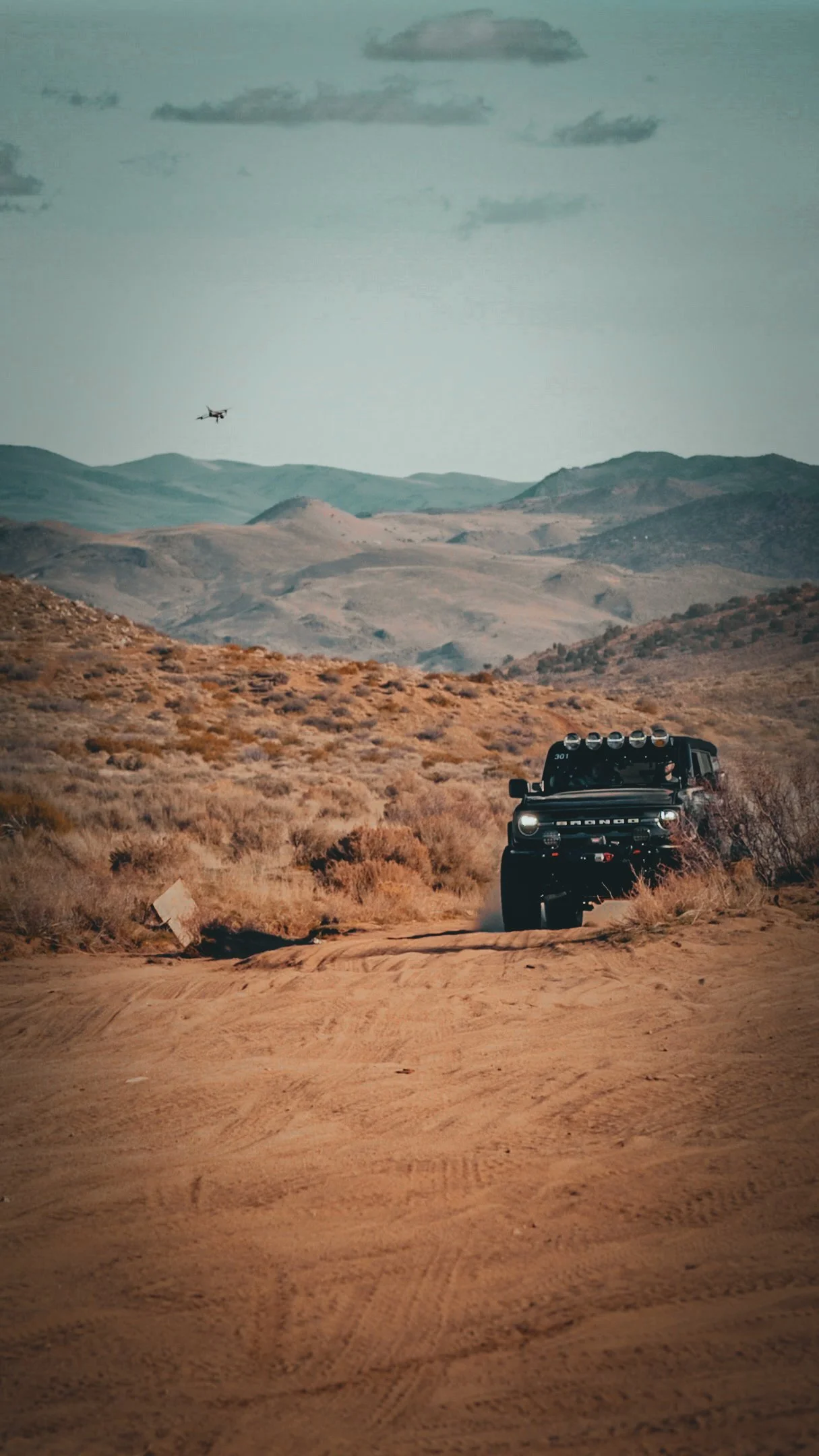 A blue Ford Bronco driving on a dirt road in a desert landscape with mountains in the background, cloudy sky, and a drone flying overhead.