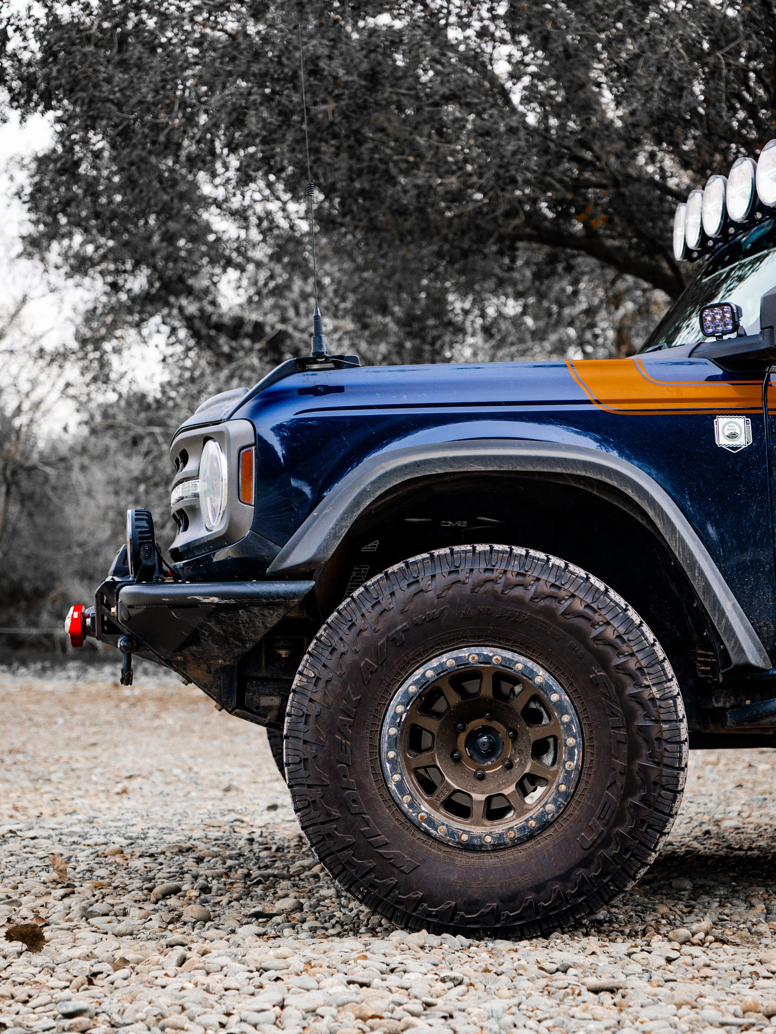 Close-up of a blue off-road vehicle's front right side, with large rugged tire, additional lights mounted on the roof, and a sticker on the door, parked on a gravel surface with trees in the background.