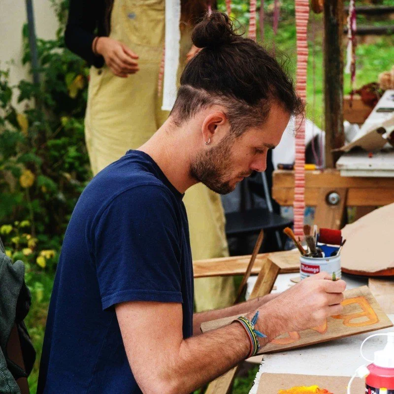 Jeune homme avec une barbe et avec un chignon, assis à une table en train de peindre ou dessiner sur un support en bois, dans un environnement en plein air, avec diverses fournitures artistiques autour.