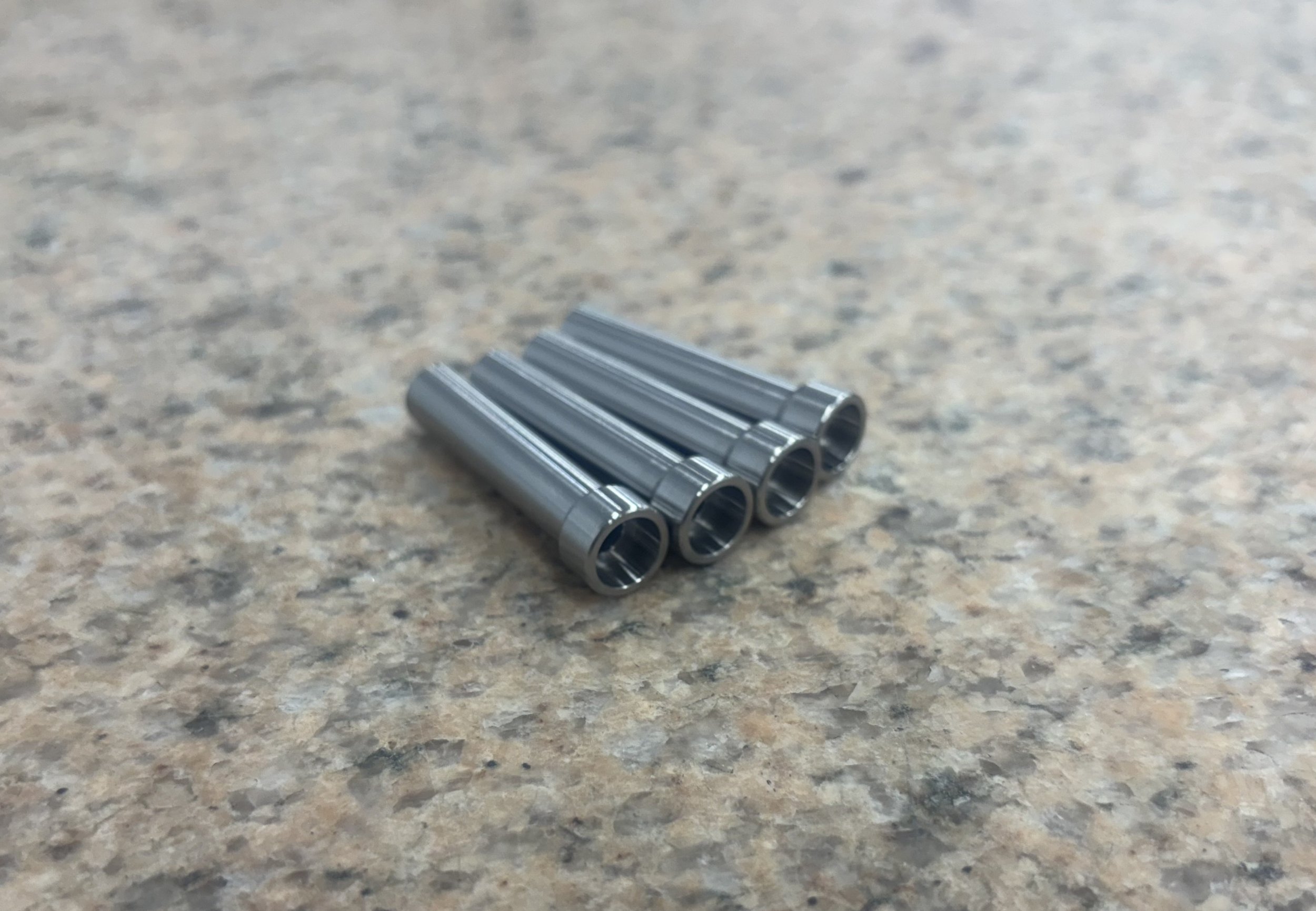 Four small metal cylindrical objects, possibly bearings or sleeves, placed on a speckled surface.