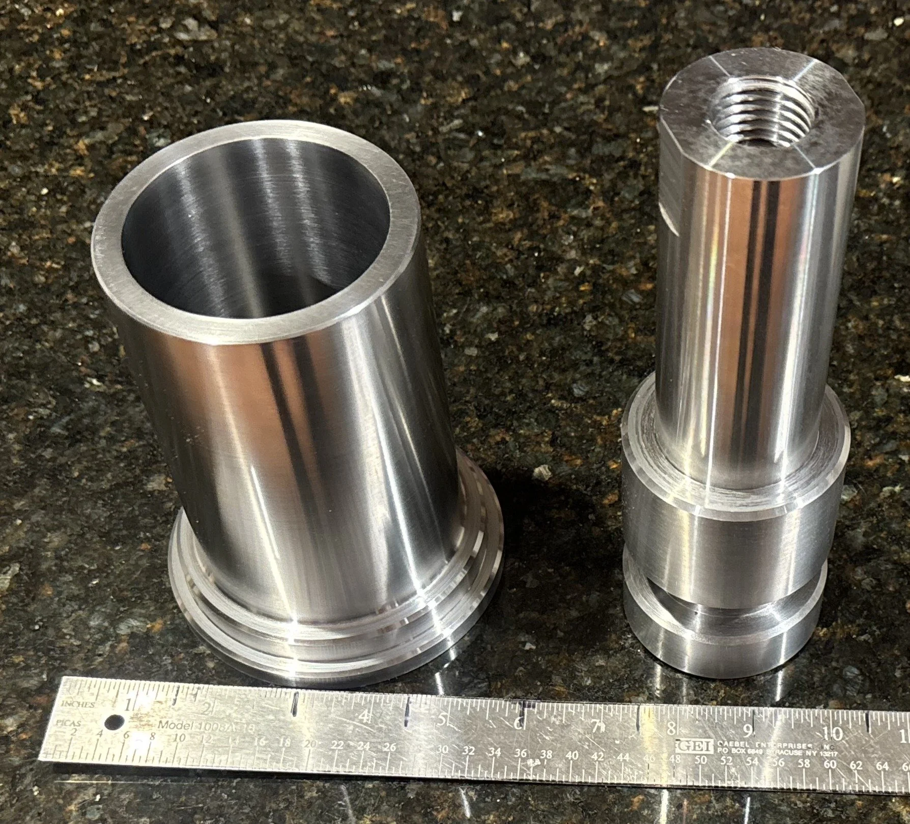 Two machined metal cylindrical parts with flanged bases and threaded internal holes on a dark granite countertop, with a ruler in inches in front for scale.