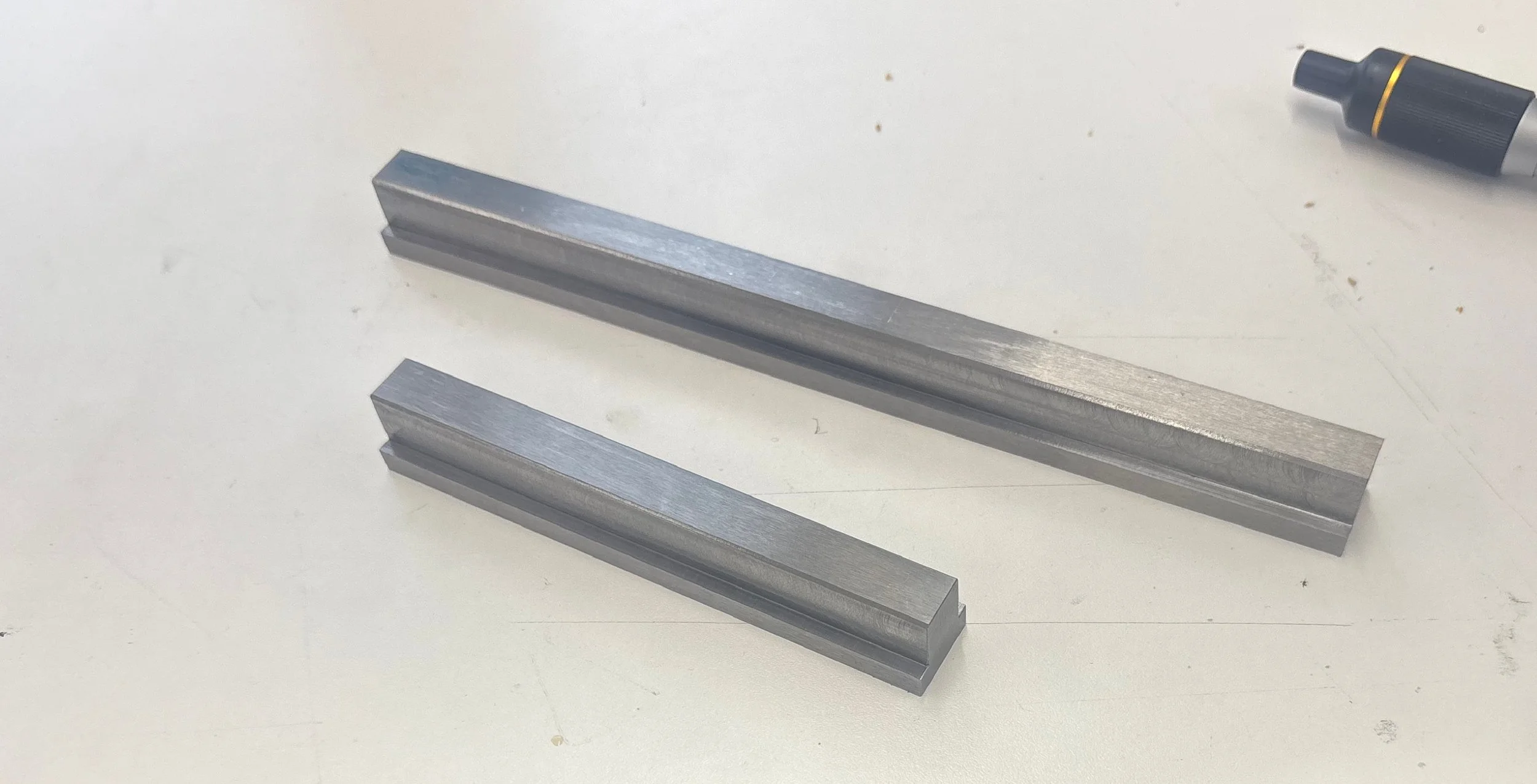 Two metallic linear objects resembling rails or bars and a black marker pen with a gold ring on the cap are placed on a white surface.