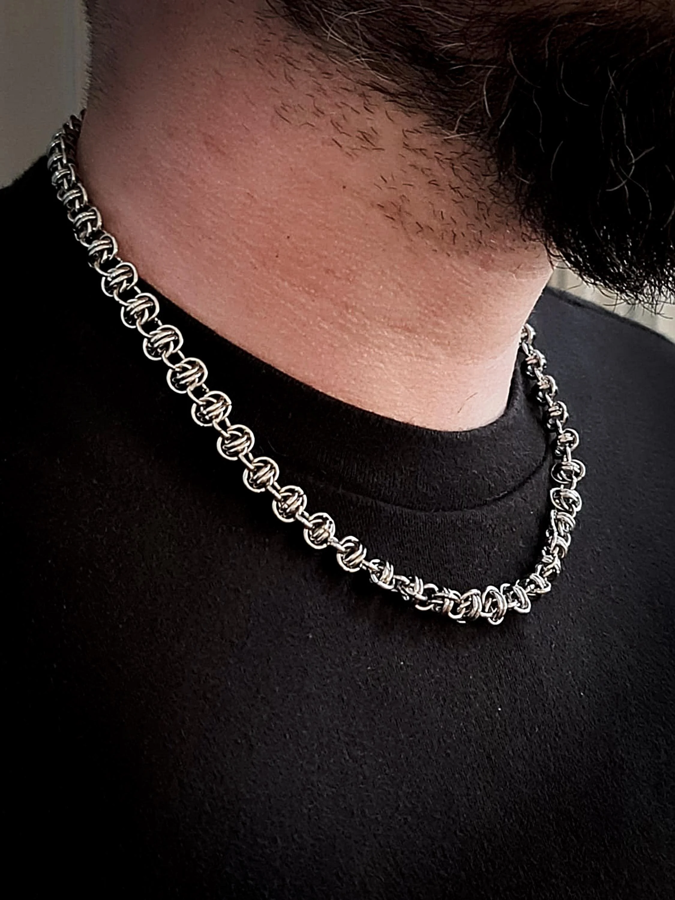 Close-up photo of a person's neck wearing a silver chain necklace, black shirt, and dark facial hair.