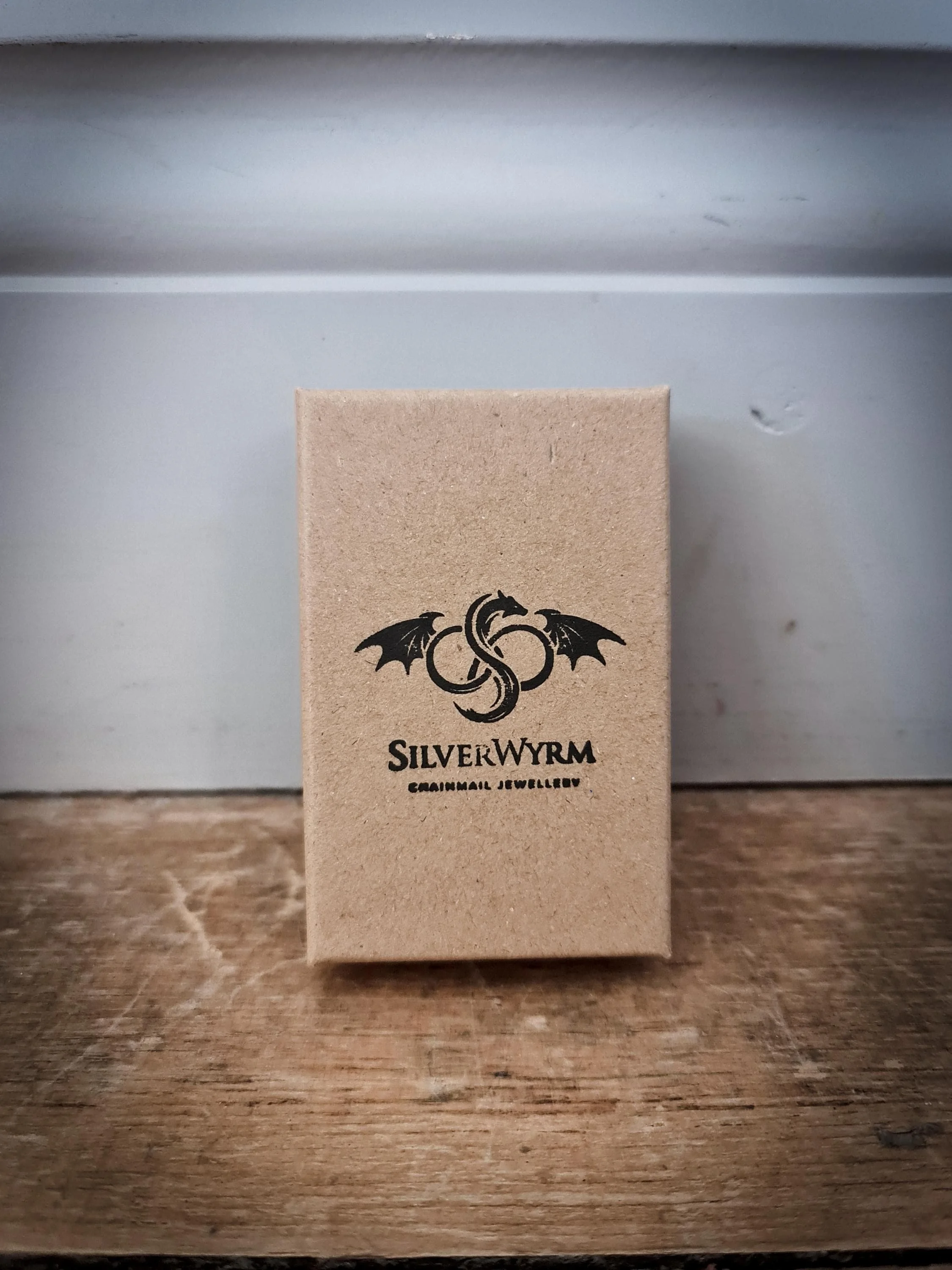 Small cardboard box with a black dragon emblem and the text 'Silverwyrm Chainmail Jewellery' printed on it, placed on a wooden surface.