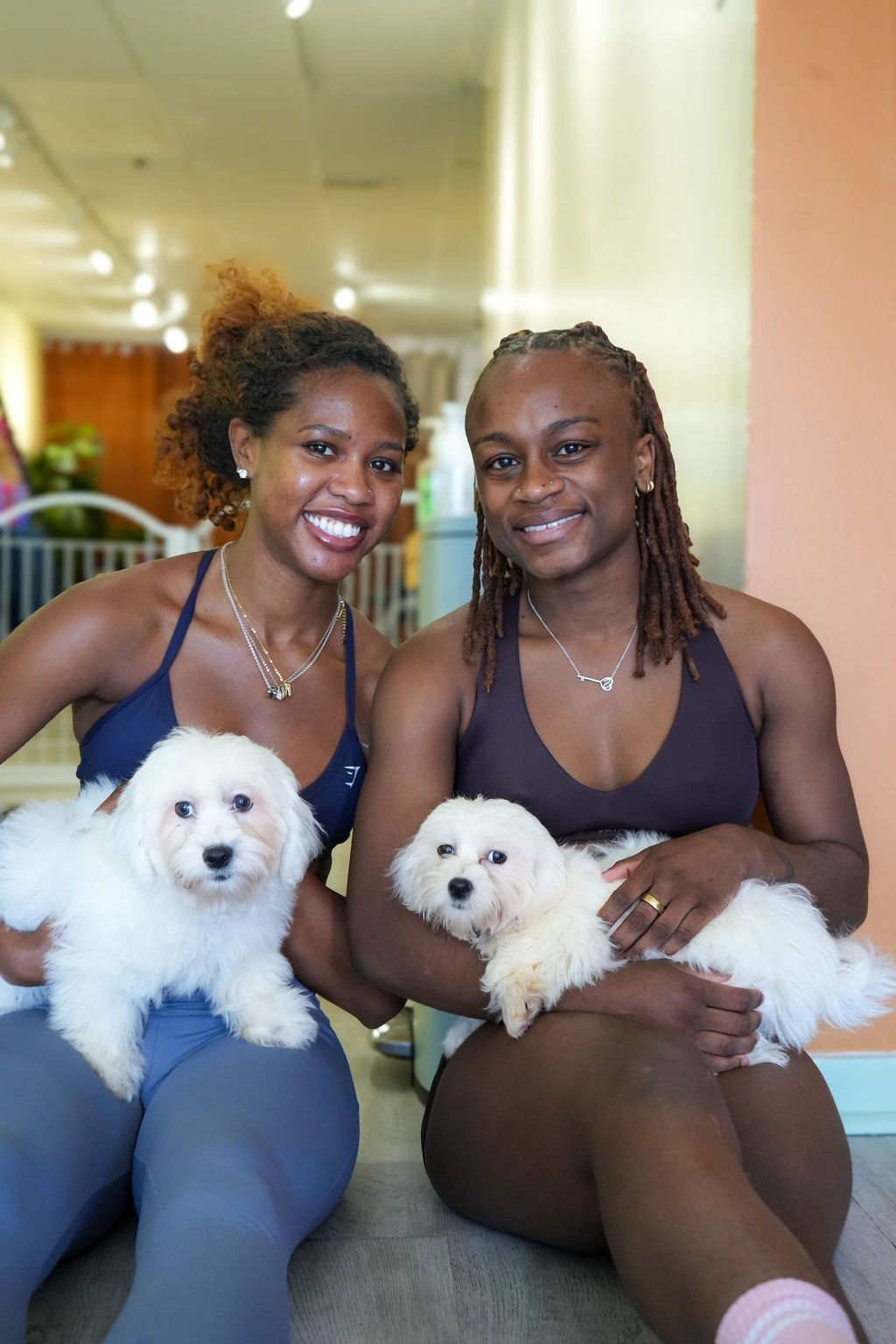 Two women sitting indoors, each holding a fluffy white puppy, smiling at the camera.