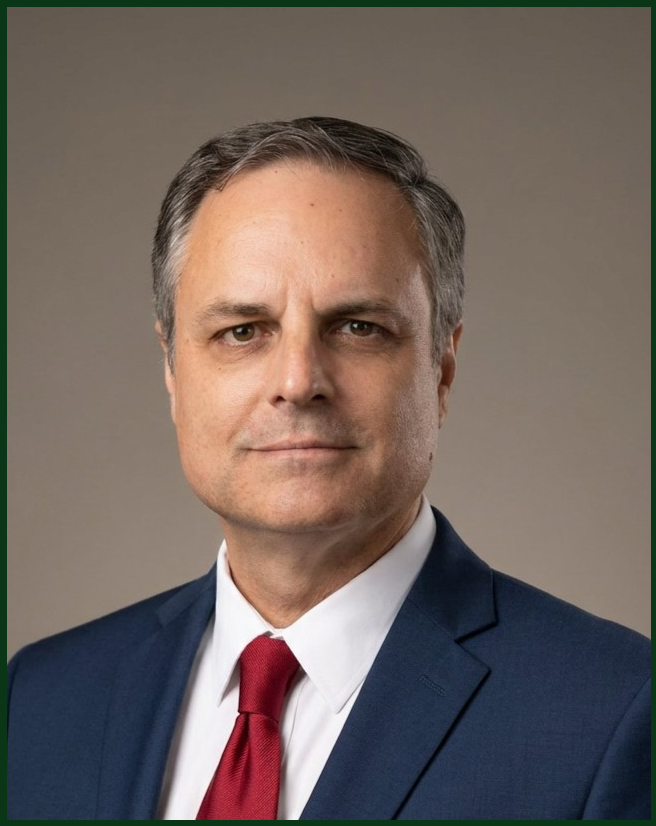A professional headshot of a middle-aged man with short, gray hair, wearing a navy blue suit, white shirt, and red tie, against a plain gray background.
