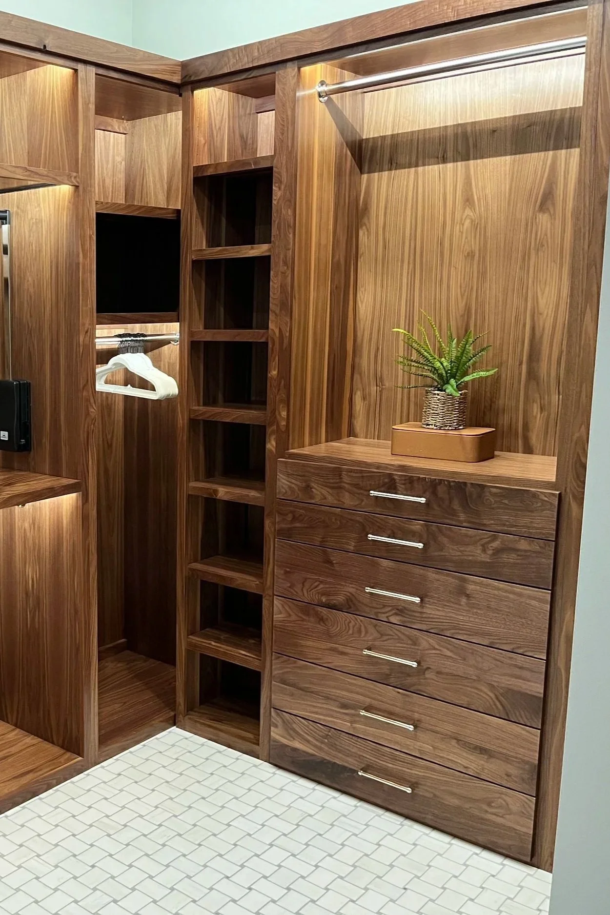 Wooden closet with open shelves and a drawer chest, decorated with a potted green plant on a small wooden tray, located in a tiled room with light mint-green walls.