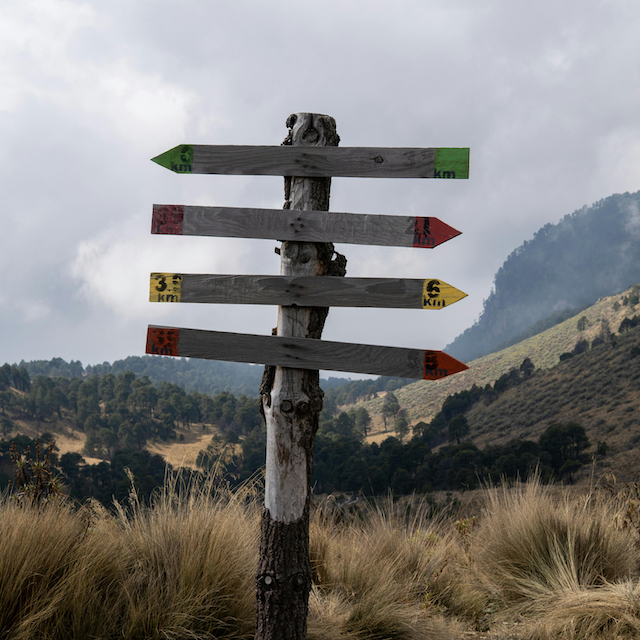 A wooden signpost with five arrows pointing in different directions, with distance markers, set against a mountainous landscape.