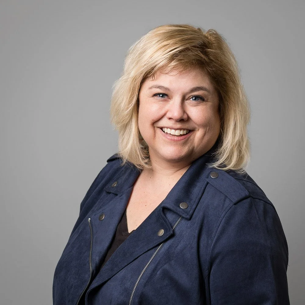 A smiling woman with shoulder-length blonde hair, wearing a dark blue jacket over a black top, posing against a plain gray background.