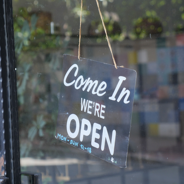 A sign hanging on a restaurant or shop window that reads, "Come In We're OPEN," indicating that the establishment is open for business.