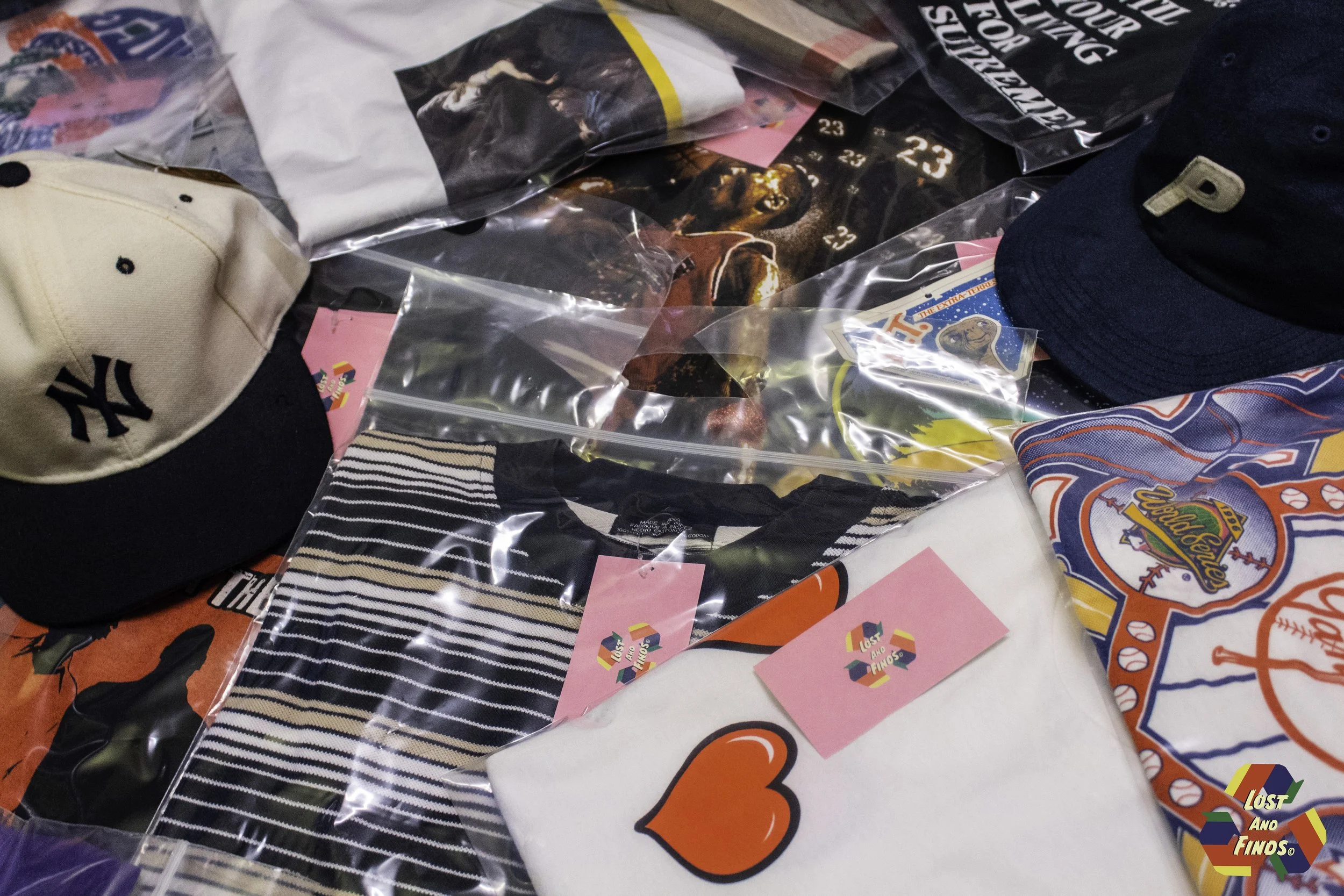 Various baseball caps, including a beige and navy Yankees cap, a white cap with red and blue embroidery, and a navy cap, are scattered on a table along with plastic bags containing different items, some with pink tags. There are also some posters or printed materials, including one with a heart symbol and another with Superzings toy packaging.
