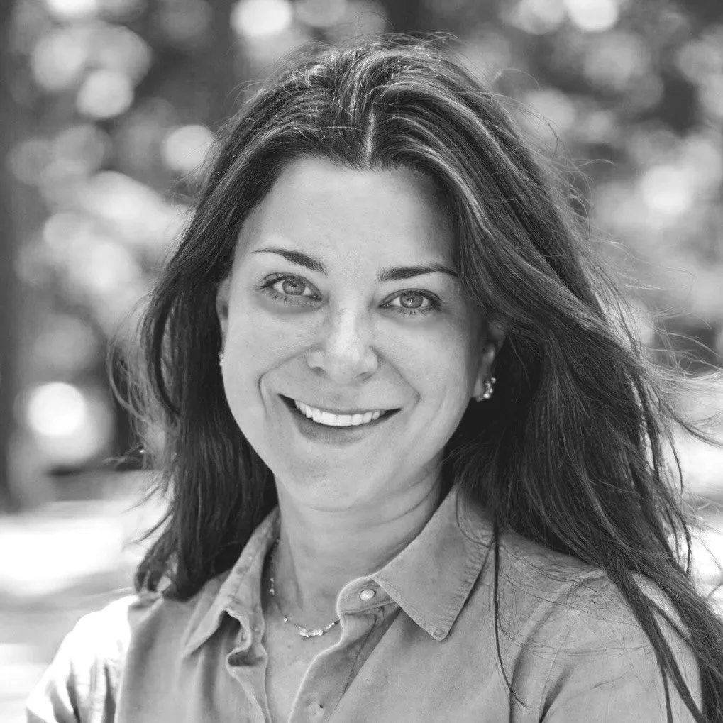 Black and white portrait of a smiling woman with long, dark hair, wearing earrings and a collared shirt, outdoors.