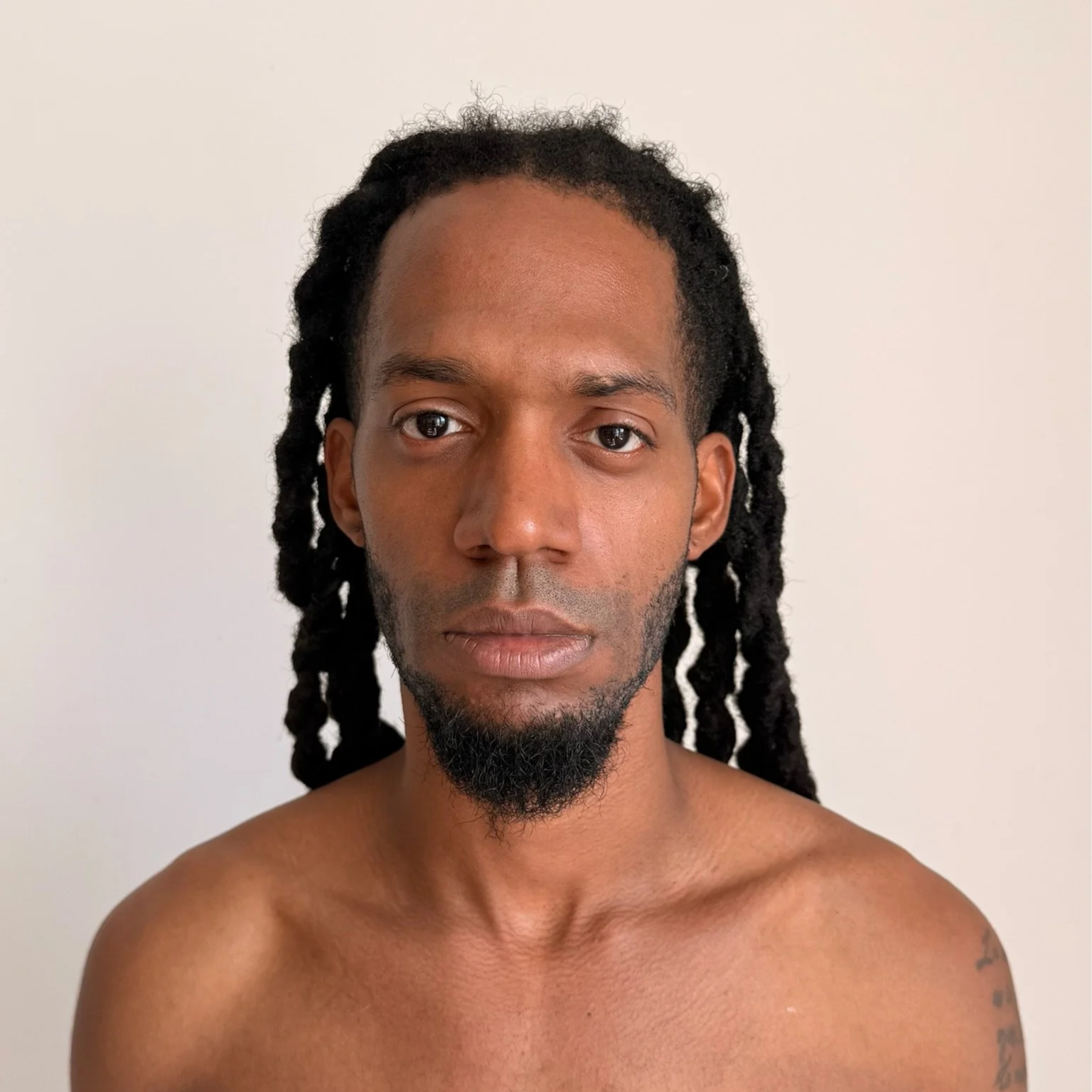 Portrait of a shirtless man with long dreadlocks and a short beard, standing against a plain white background.