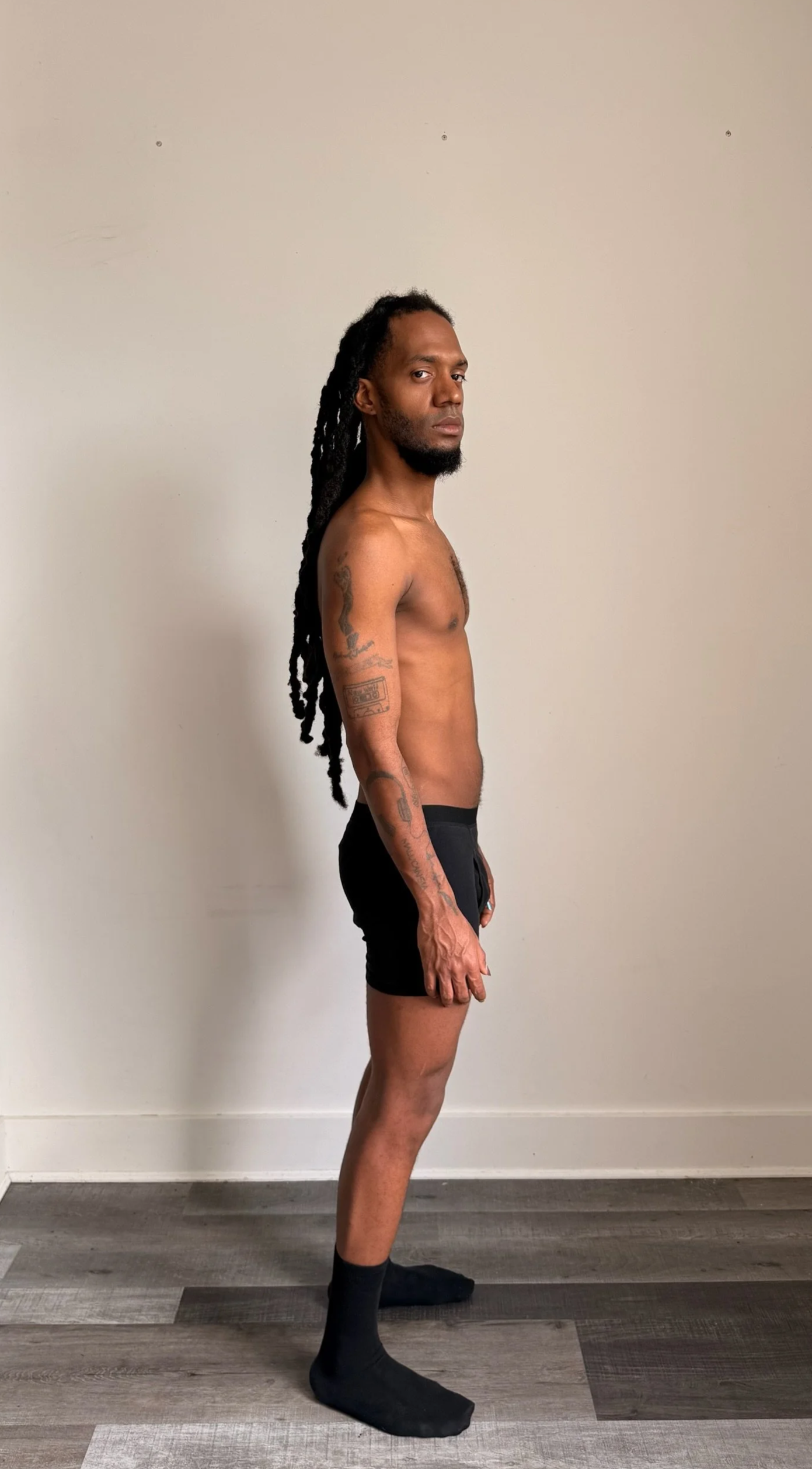 A man with dreadlocks and tattoos on his arms, standing in profile against a plain white wall, wearing black shorts and black socks.