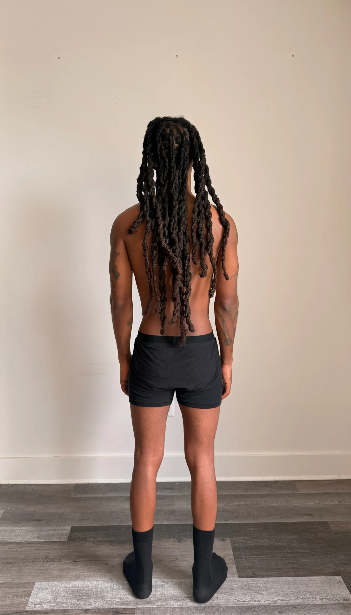 A person with long dreadlocks, shirtless, wearing black shorts and black socks standing with their back to the camera against a plain white wall.