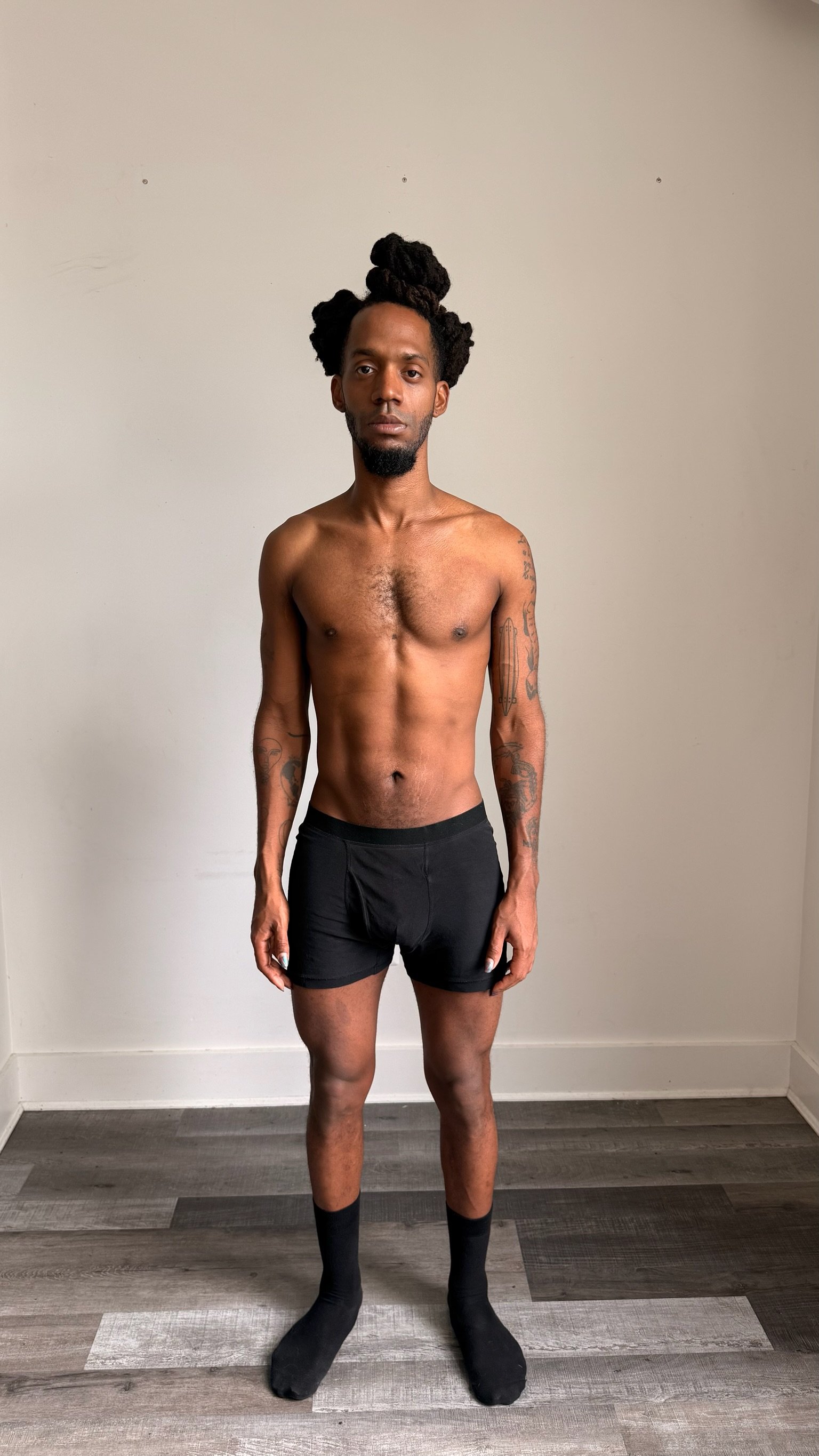 A shirtless man with dreadlocks and tattoos on both arms stands in front of a plain wall, wearing black shorts and black socks.