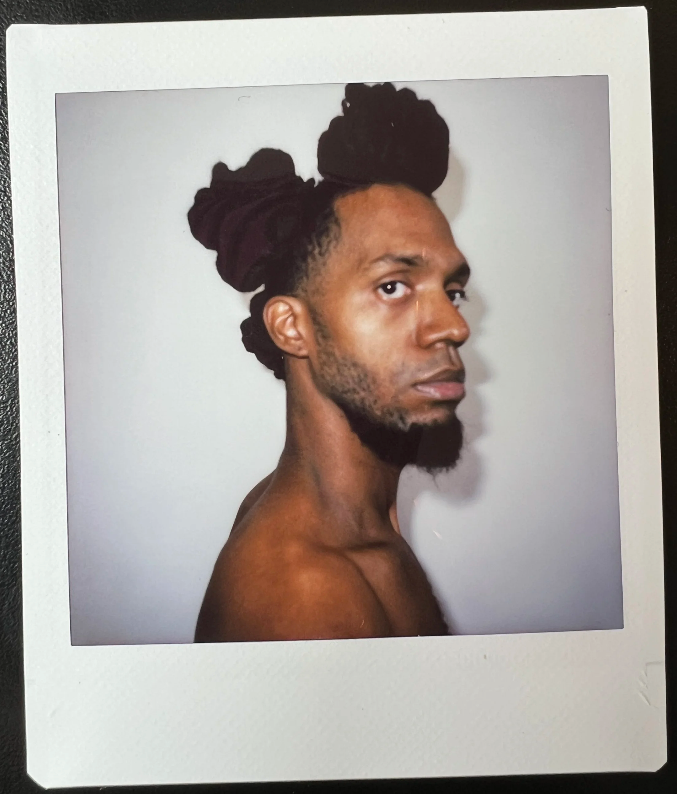 Portrait of a man with dark skin and dreadlocks styled in a high bun, looking to the side, photographed on a white background.