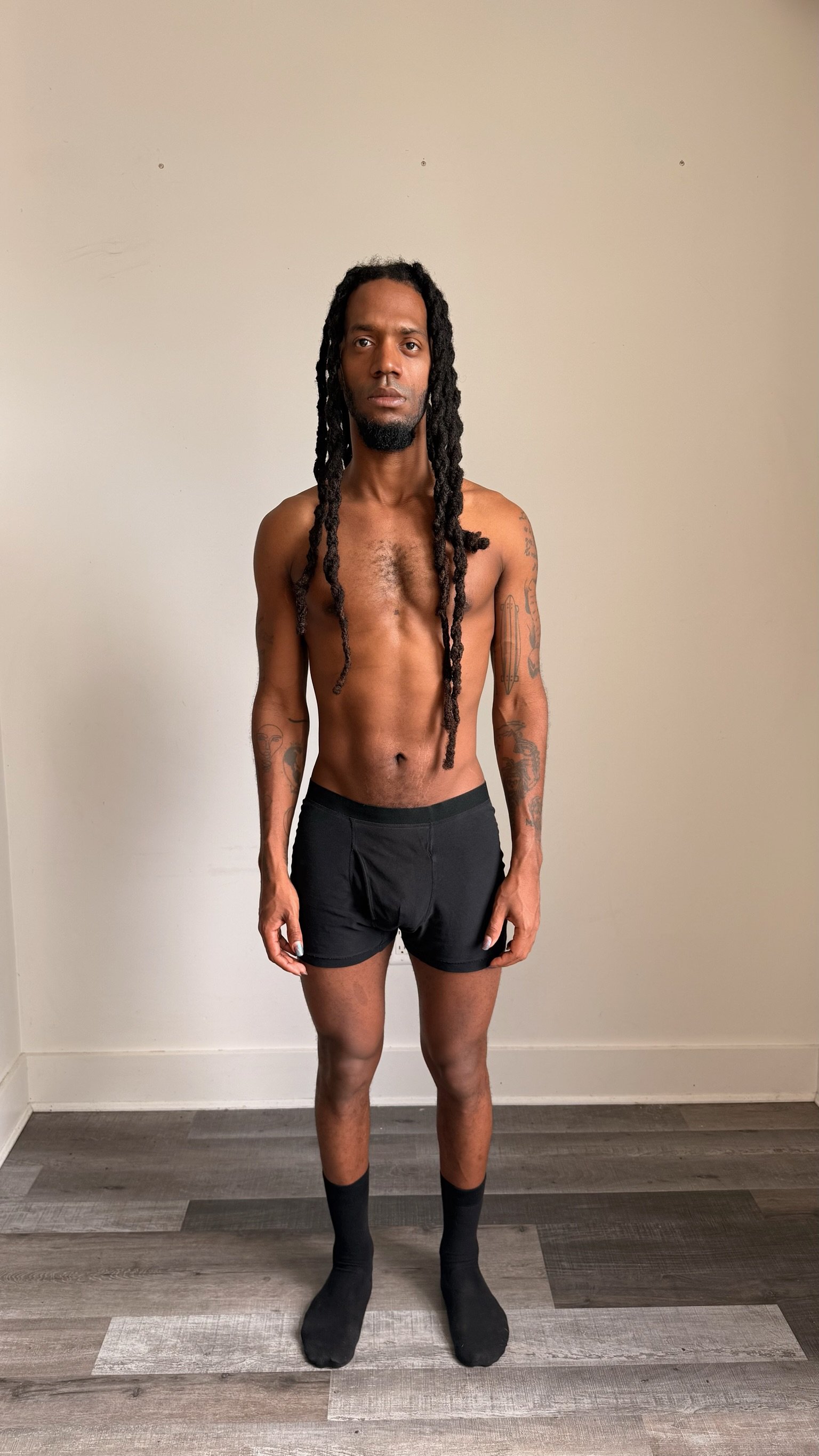 A shirtless man with long dreadlocks and tattoos standing against a plain white wall.
