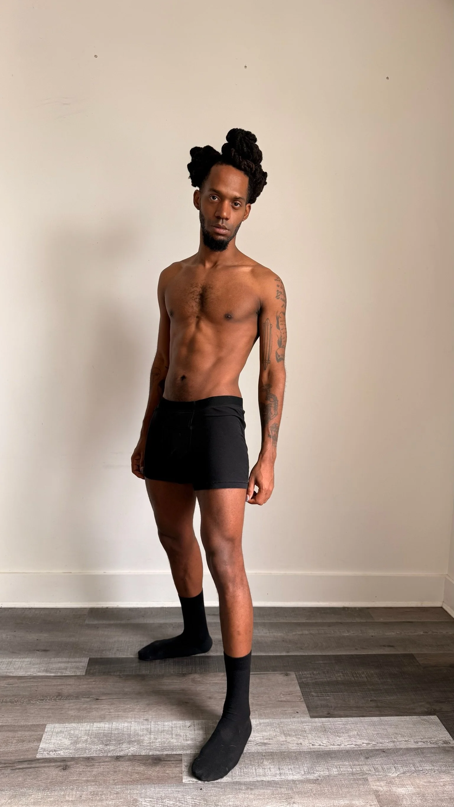 A fit topless man with black shorts and long black socks standing against a plain white wall with a wood floor.