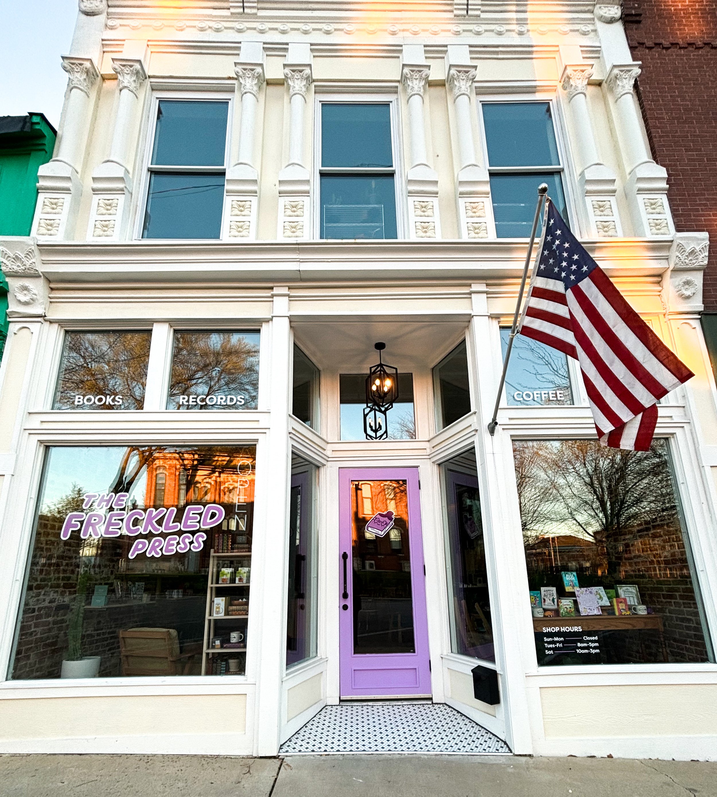 The entrance of a bookstore called 'The Freckled Bookpress' with a purple door, display windows showing books and merchandise, and an American flag hanging outside. Shop hours are posted on the window.