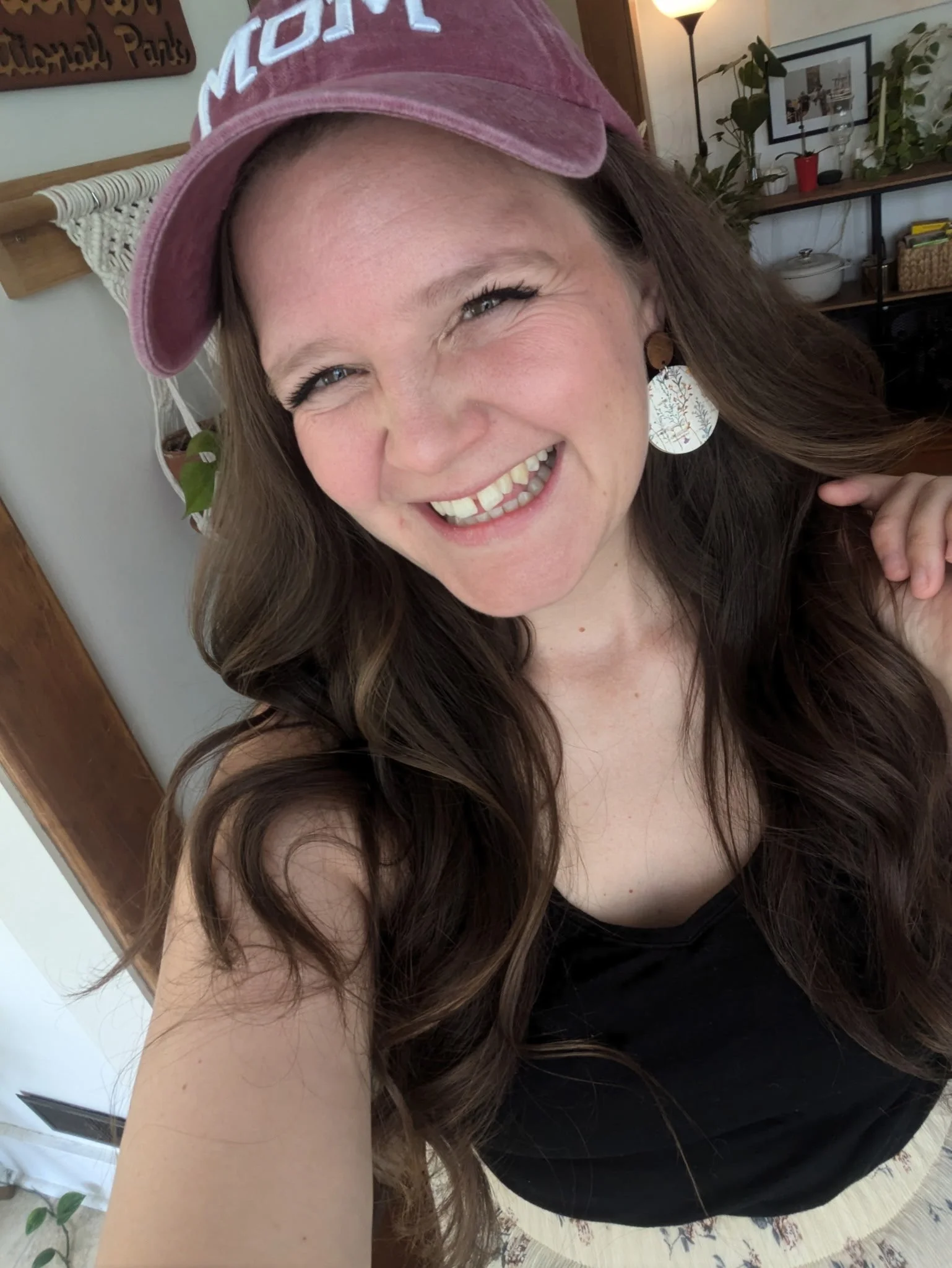 A woman with long brown hair smiling while taking a selfie indoors. She is wearing a pink velvet baseball cap and a black top. In the background, there is a lamp, a plant, framed pictures, and a bookshelf.