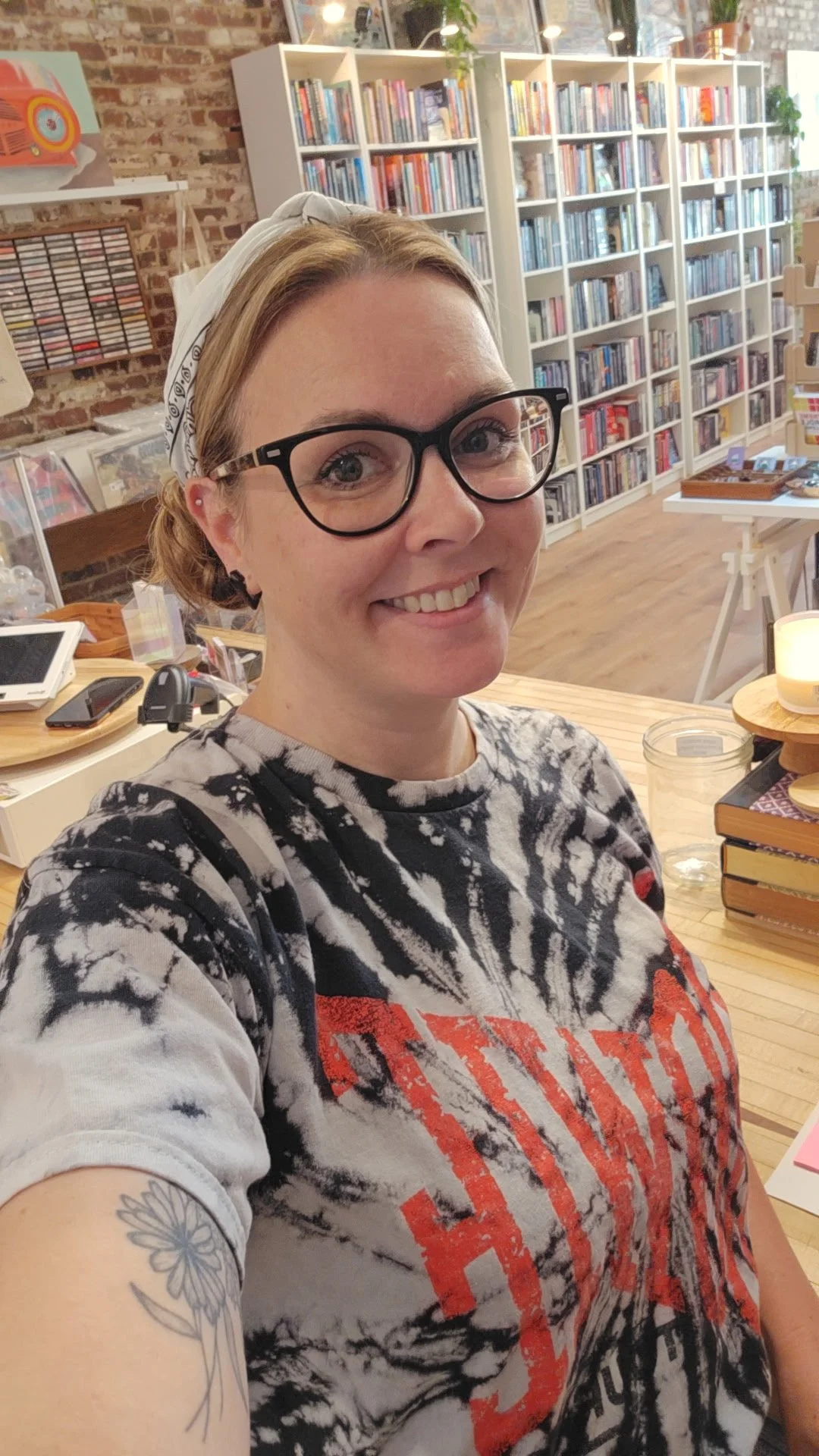 A woman with glasses and a bandana taking a selfie in a bookstore or library with bookshelves and wooden furniture in the background.