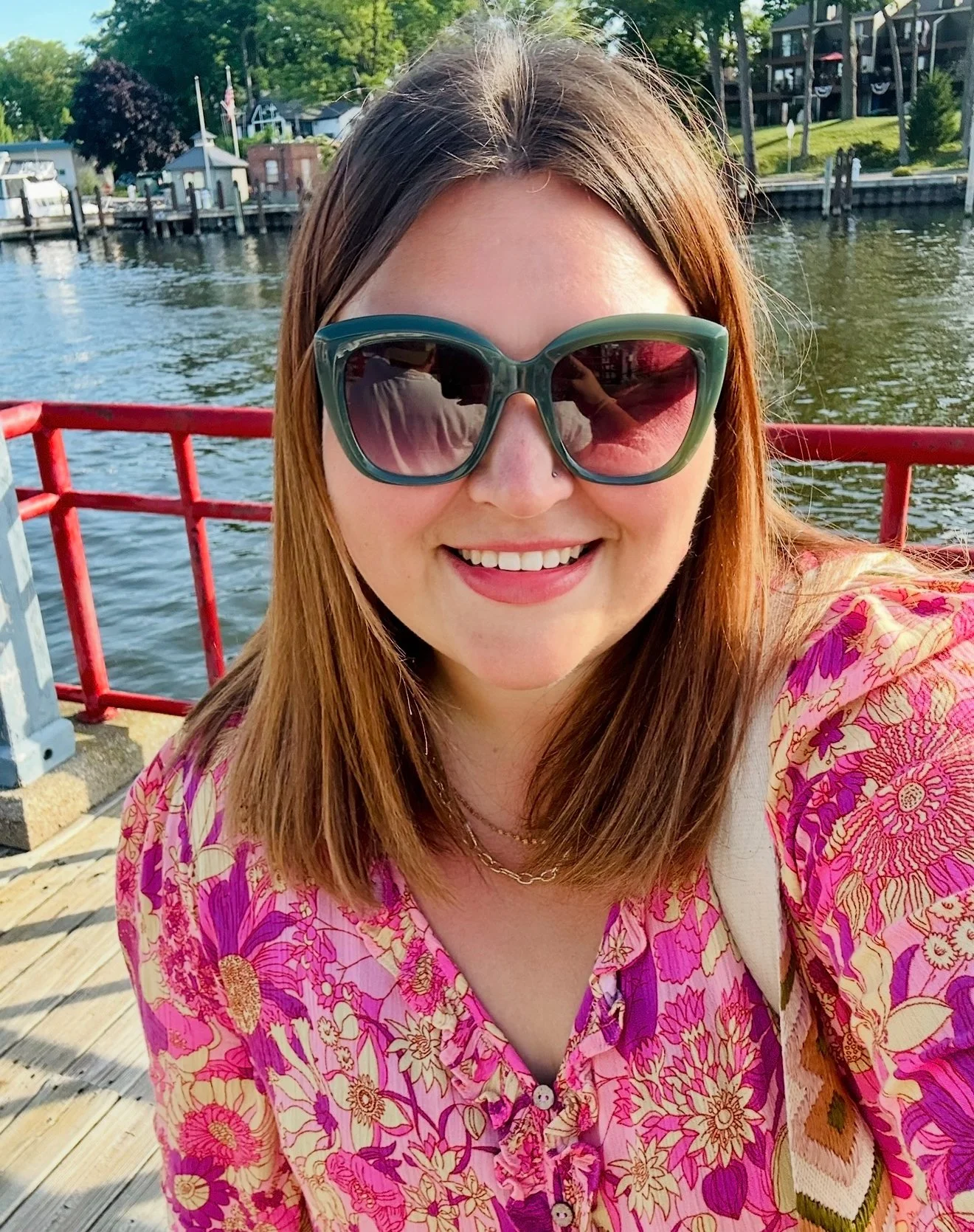 A woman with long red hair wearing large sunglasses and a pink floral shirt is smiling for a selfie outdoors near a body of water with docks and boats, and green trees in the background.