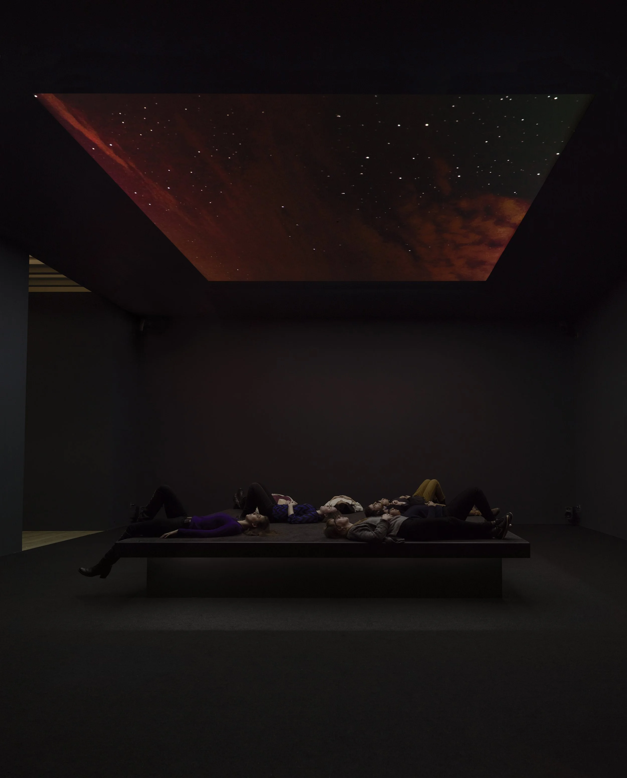 People lying on a black platform in a dark room with a galaxy and starry sky projected on the ceiling. ASTRO NOISE exhibition view.