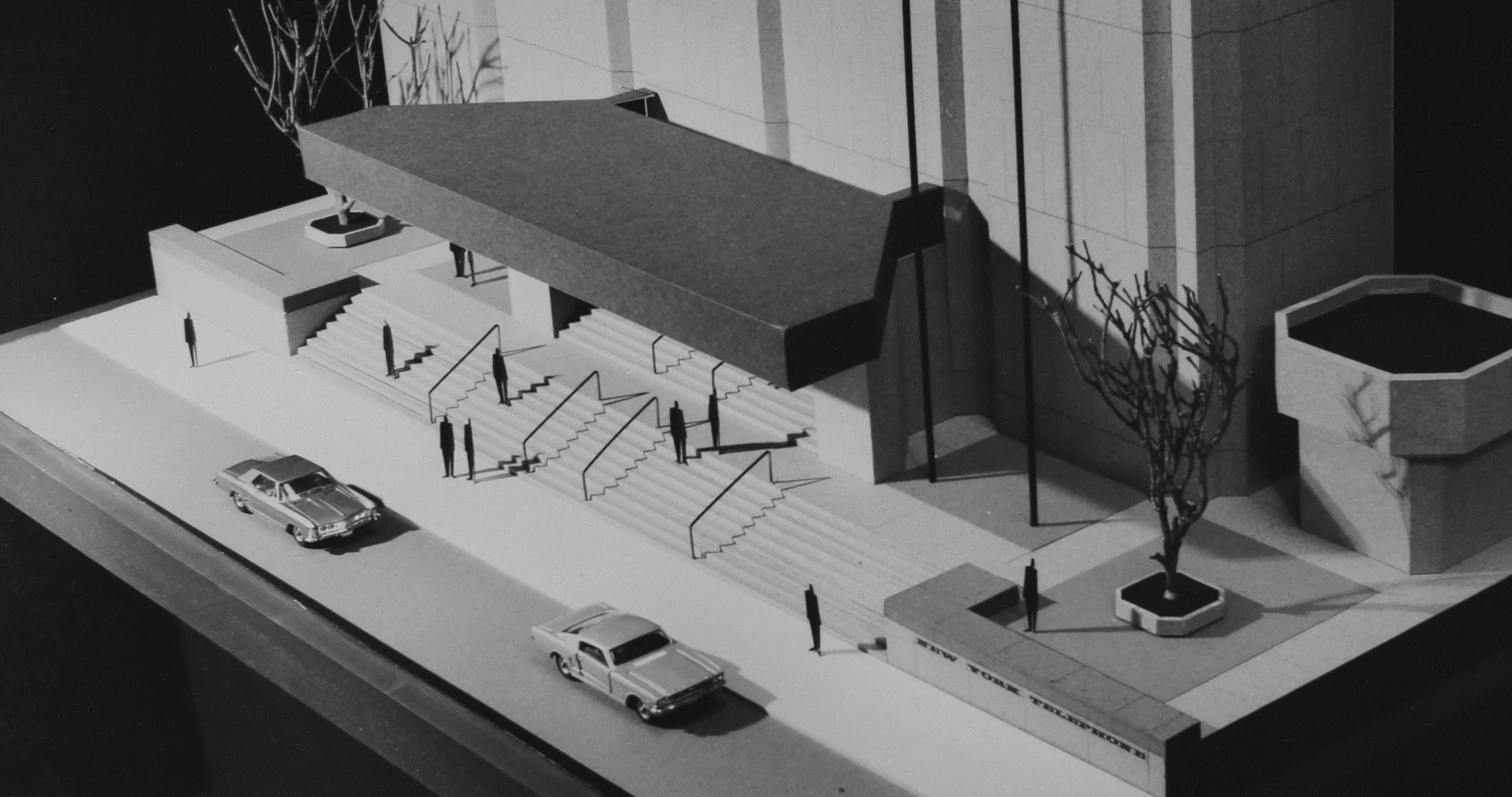 Black and white architectural model of a brutalist building with stairs leading up to the entrance, surrounded by small trees and parked cars on the street.