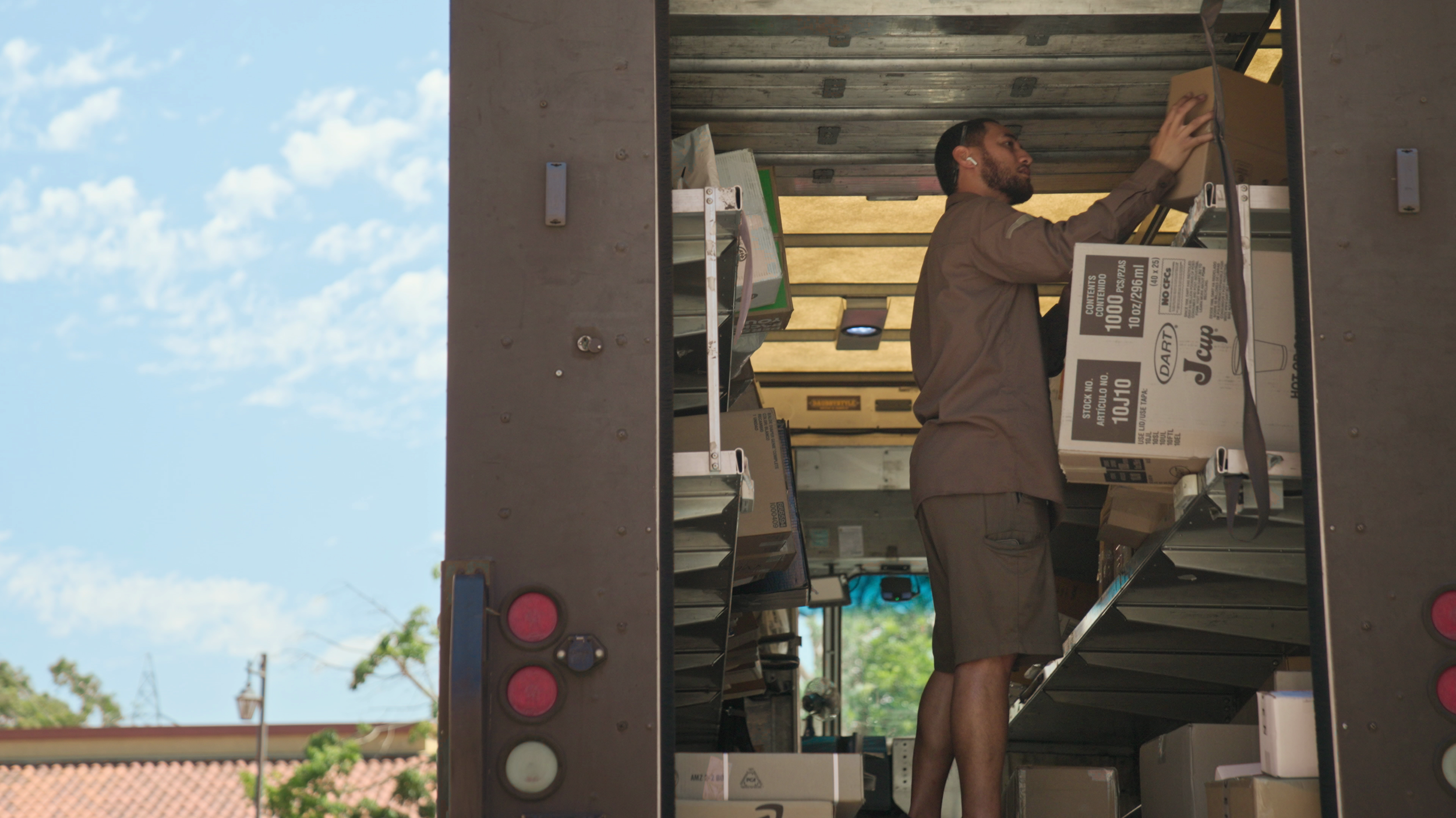A UPS driver working a delivery truck, handling a boxed item. The truck is parked outdoors under a clear sky with some clouds visible.