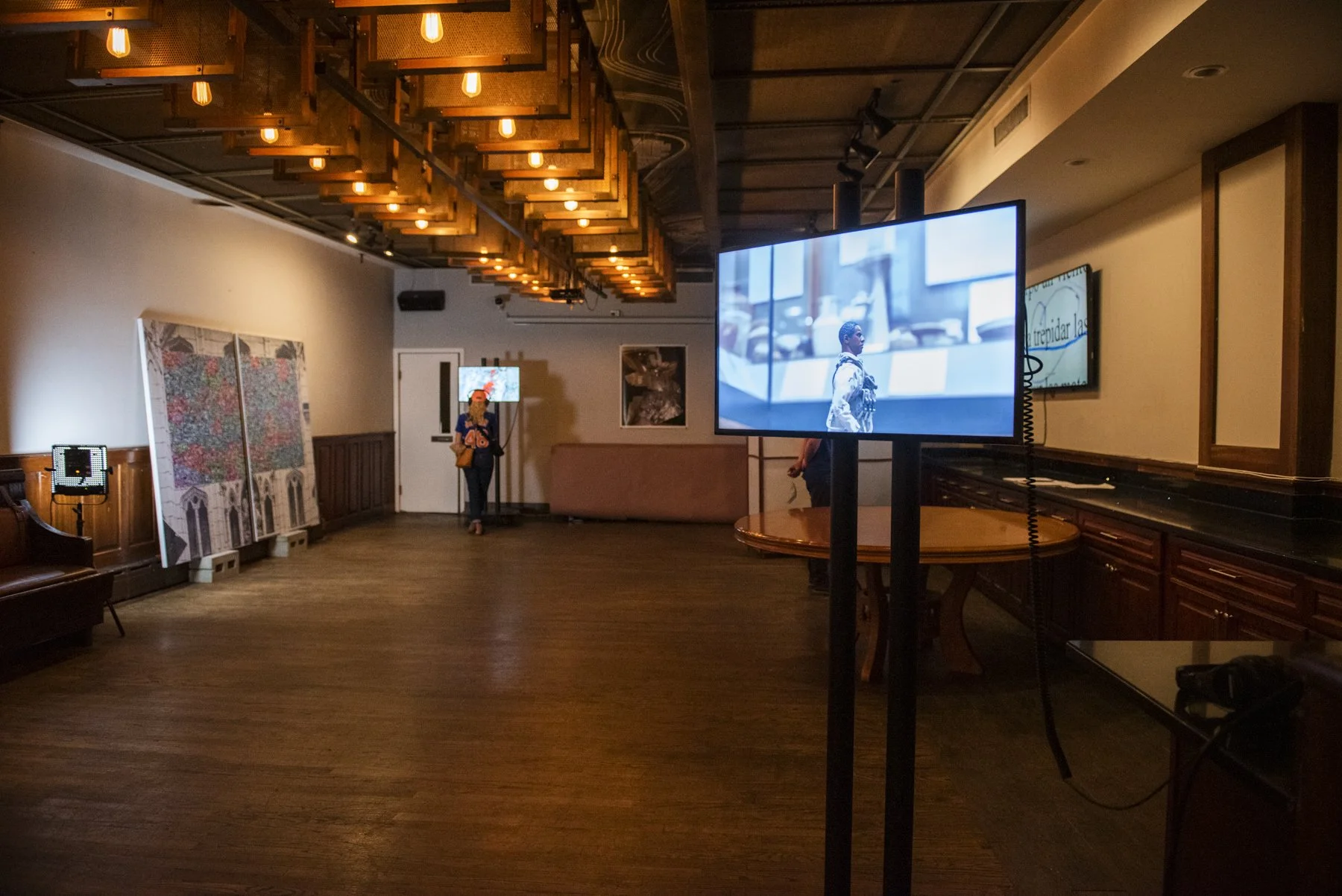 Installation view of PARALLEL CONSTRUCTION. Two people are standing in the back, various art is displayed on TV screens and mounted on walls.