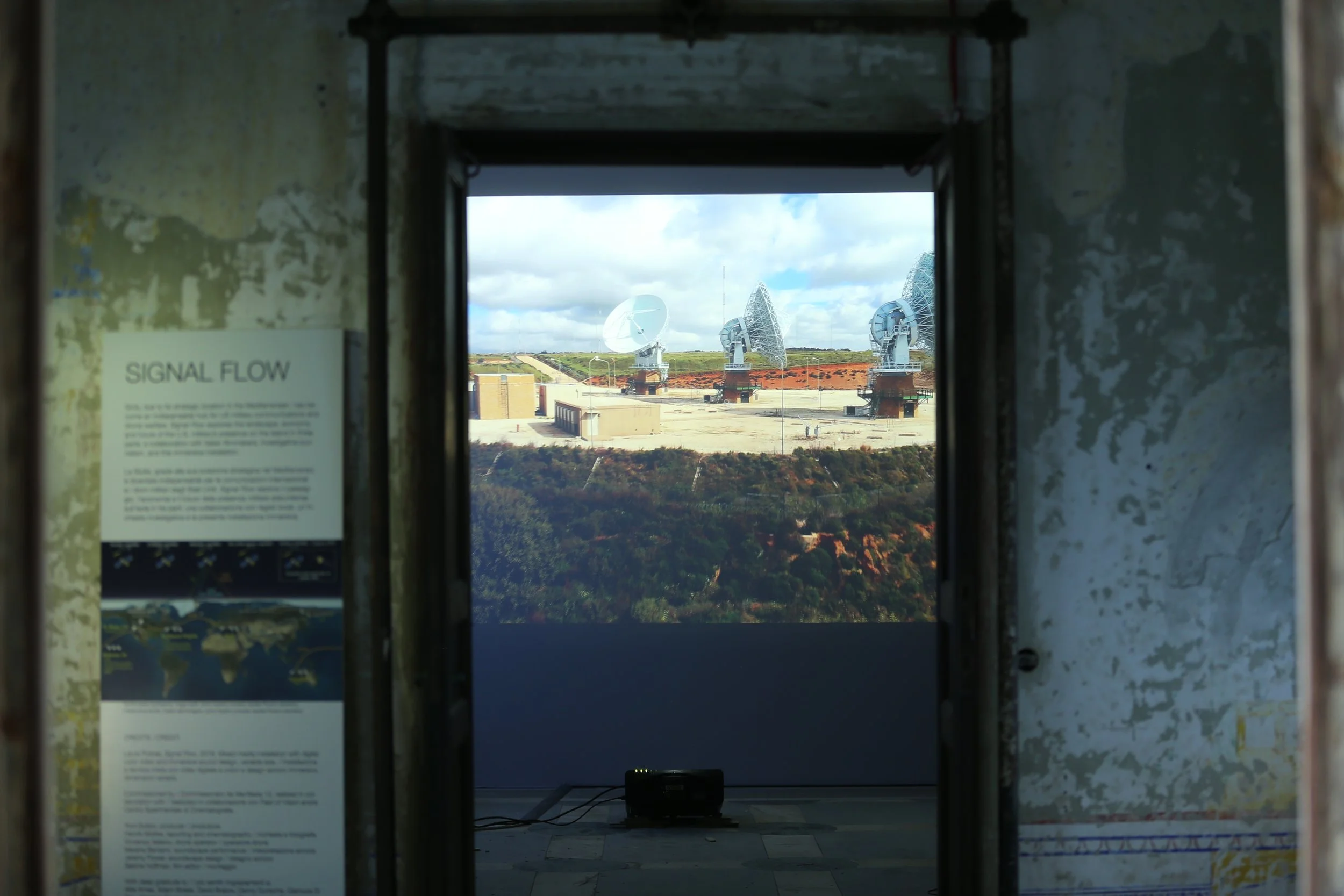 Installation view of exhibition SIGNAL FLOW, a photo through a doorway showing a film projection of satellite dishes. 