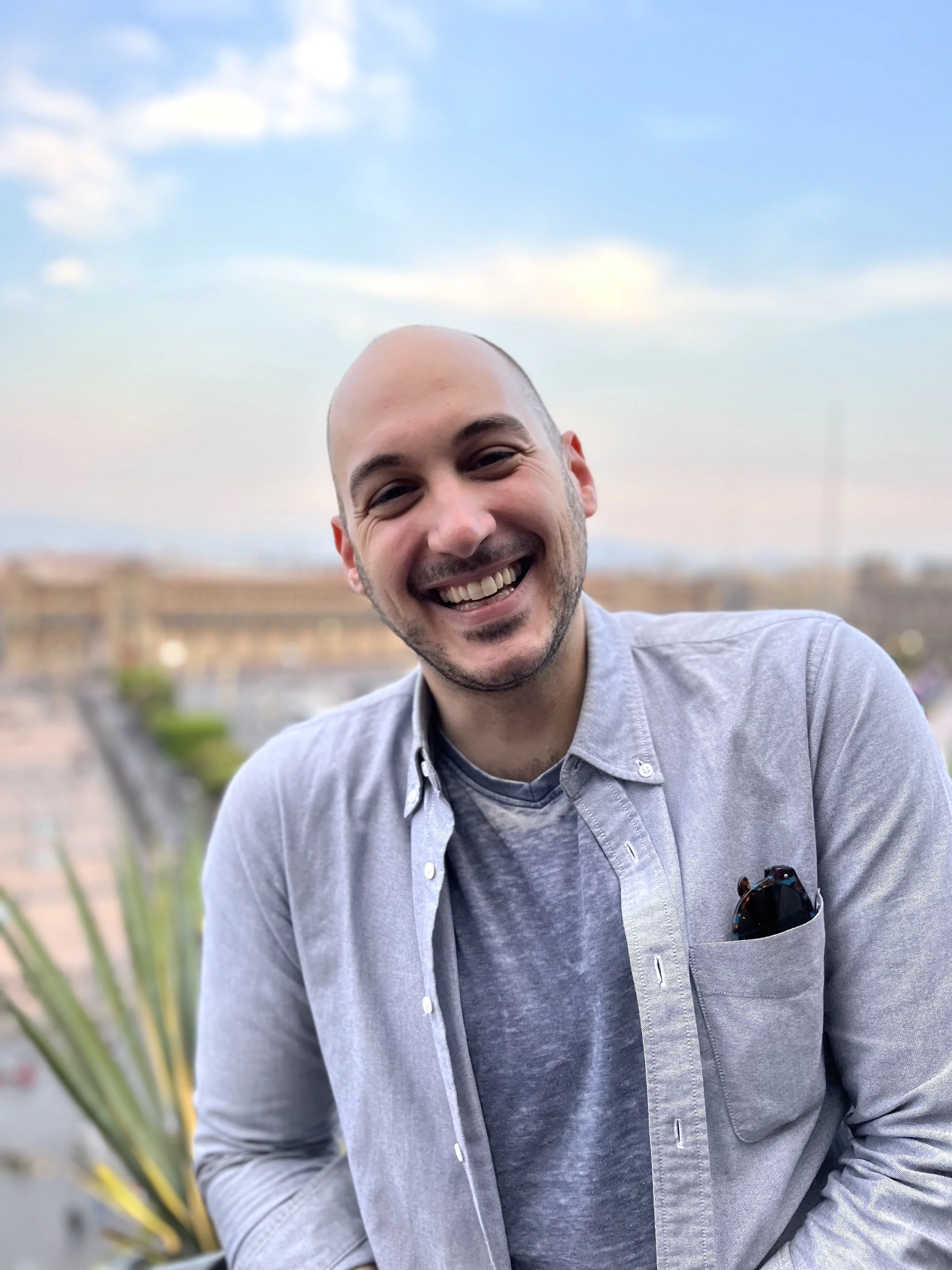 A photograph of Yoni Golijov, he is a smiling white man with a shaved head and beard, wearing a grey shirt over a t-shirt, smiling outdoors with a blurred cityscape and blue sky in the background.