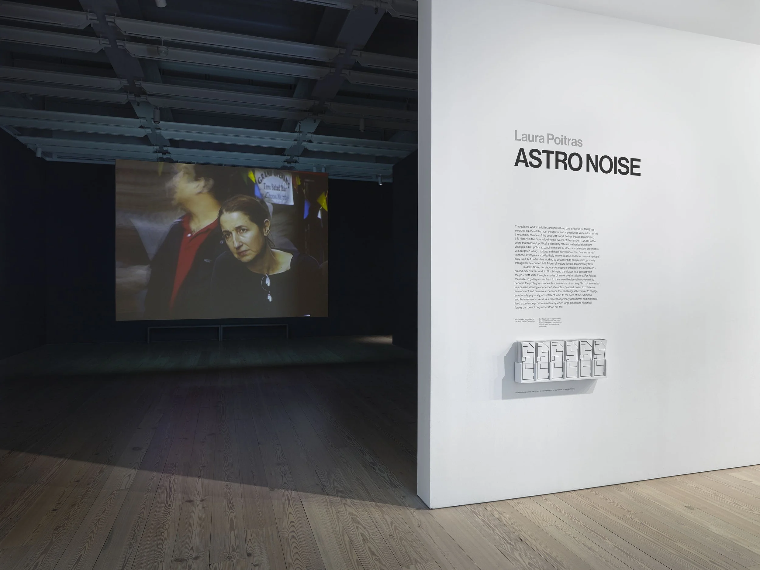 Installation view of the exhibition 'ASTRO NOISE' by Laura Poitras. To the left, there is a screen displaying a film with a woman looking into the distance.