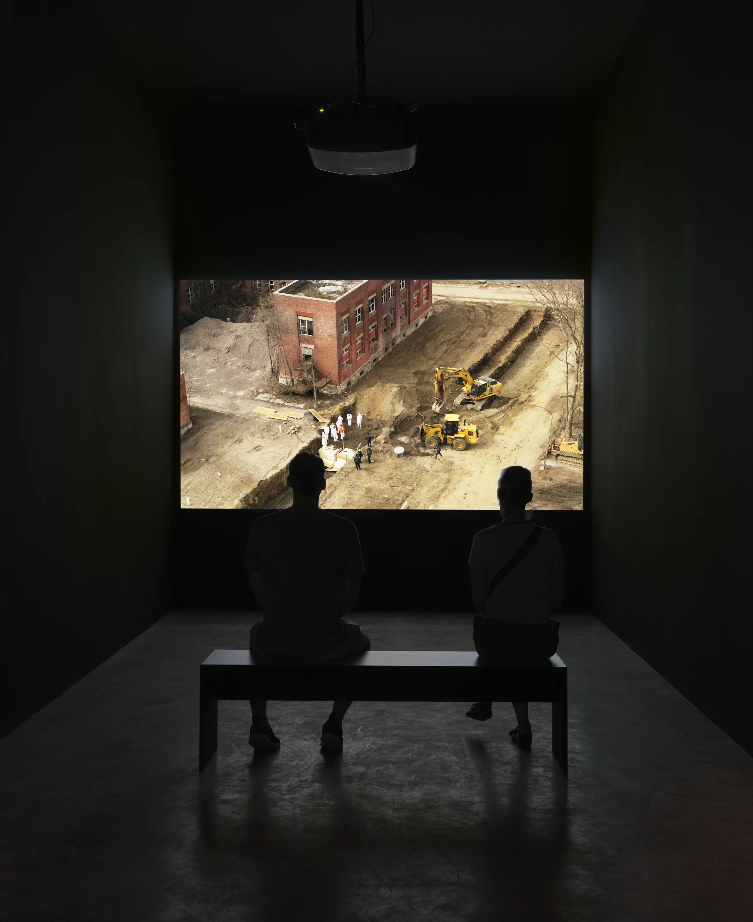 Two people sitting on a bench in a dark room watching a video of a mass burial site on a large projection. CIRCLES exhibition view.