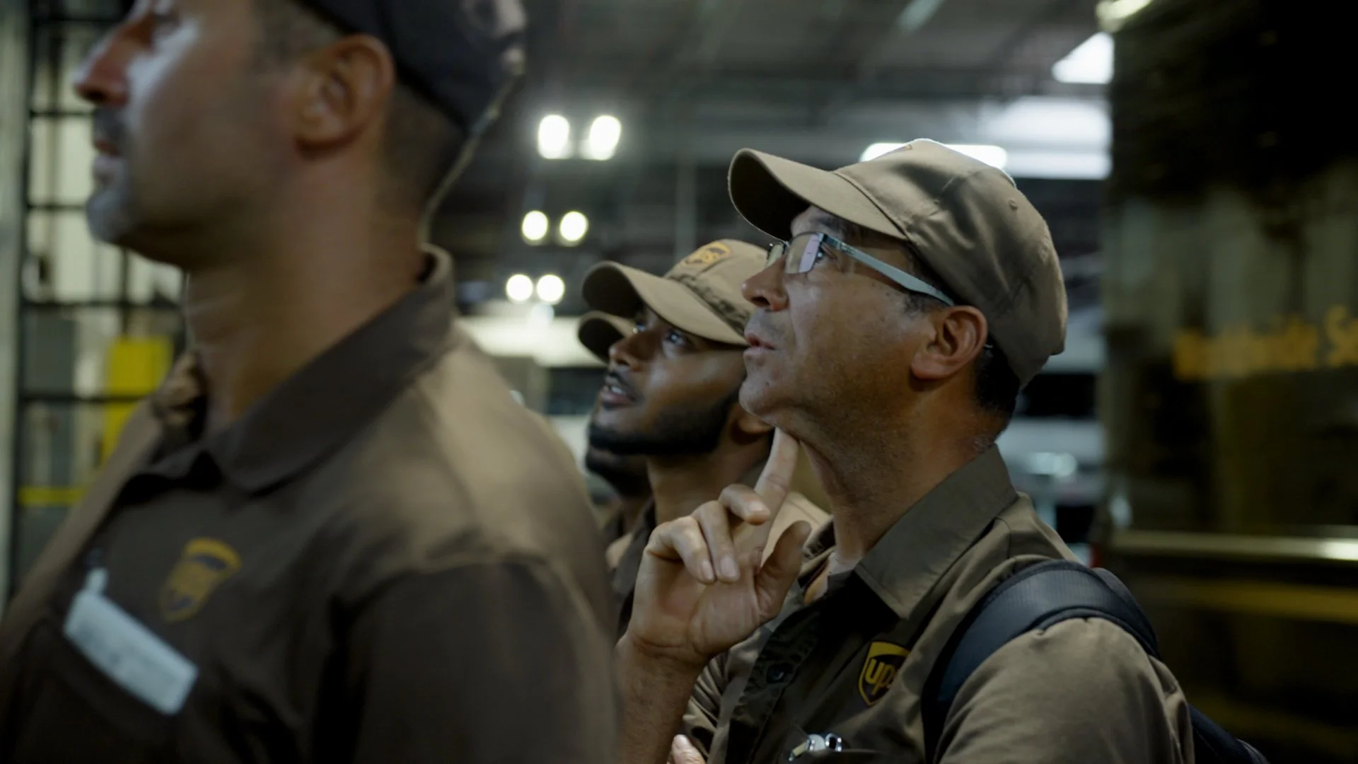 A group of UPS workers in uniform inside a warehouse, standing in a row and looking up into the distance.