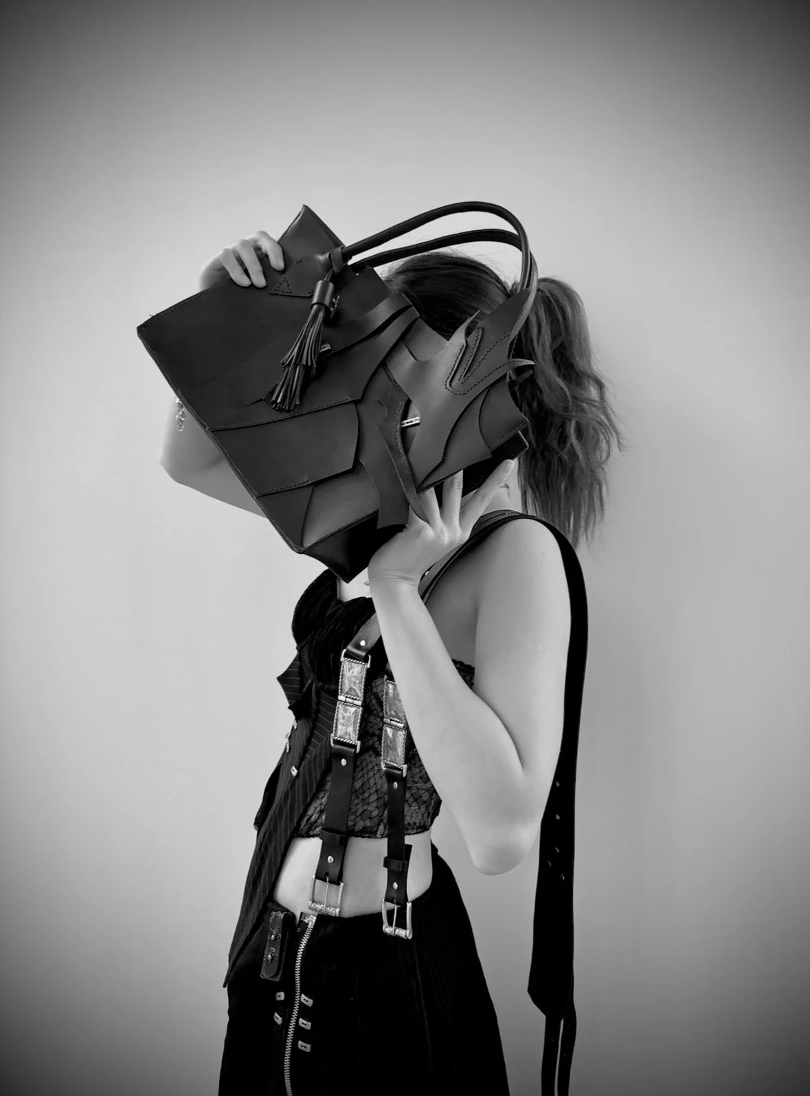 A woman with wavy hair holding a large designer handbag in front of her face, wearing a sleeveless top with straps and a zip-up skirt, against a plain background.