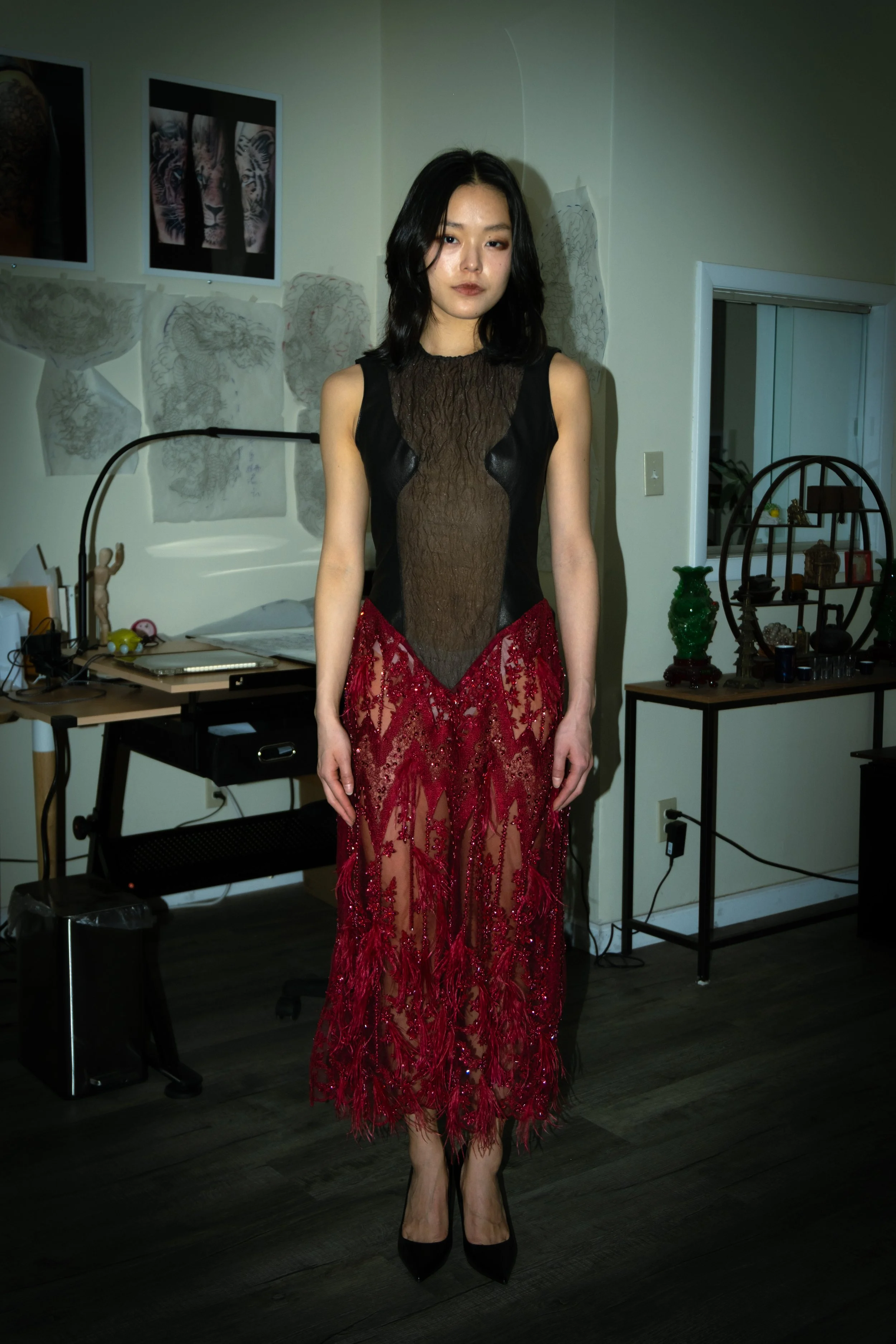 A woman with black hair wearing a black and red lace evening gown standing indoors, with artwork on the wall and furniture behind her.