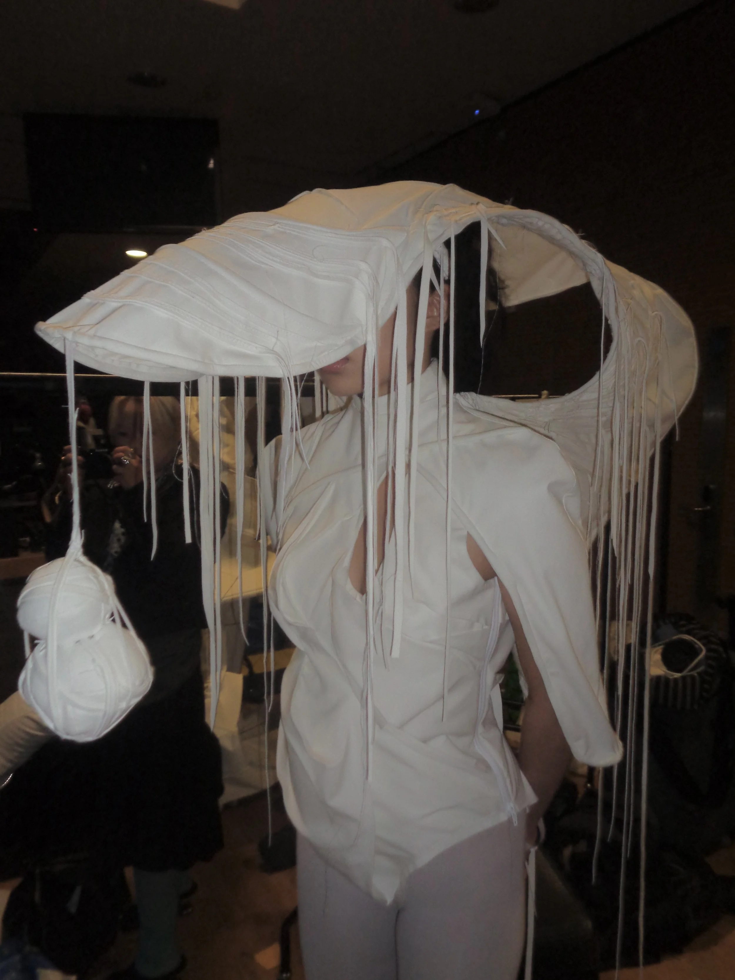 Person wearing an avant-garde white fashion piece with long hanging wires and layered fabric, partially covering their face, in a dim room with other people in the background.