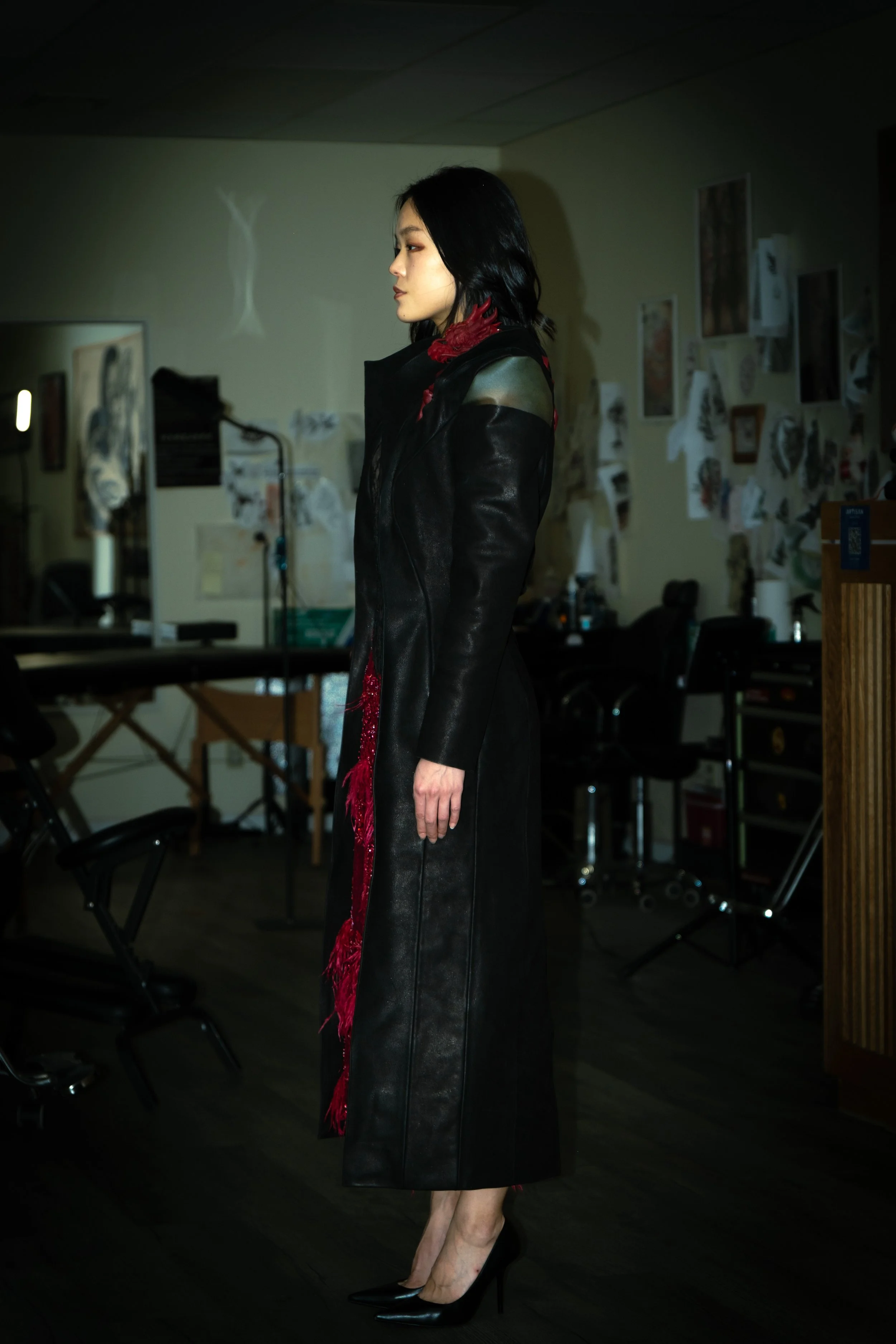 A woman with wavy black hair in a black leather coat with red accents stands in a room with artist's sketches and paintings on the wall. She is wearing black high heels.