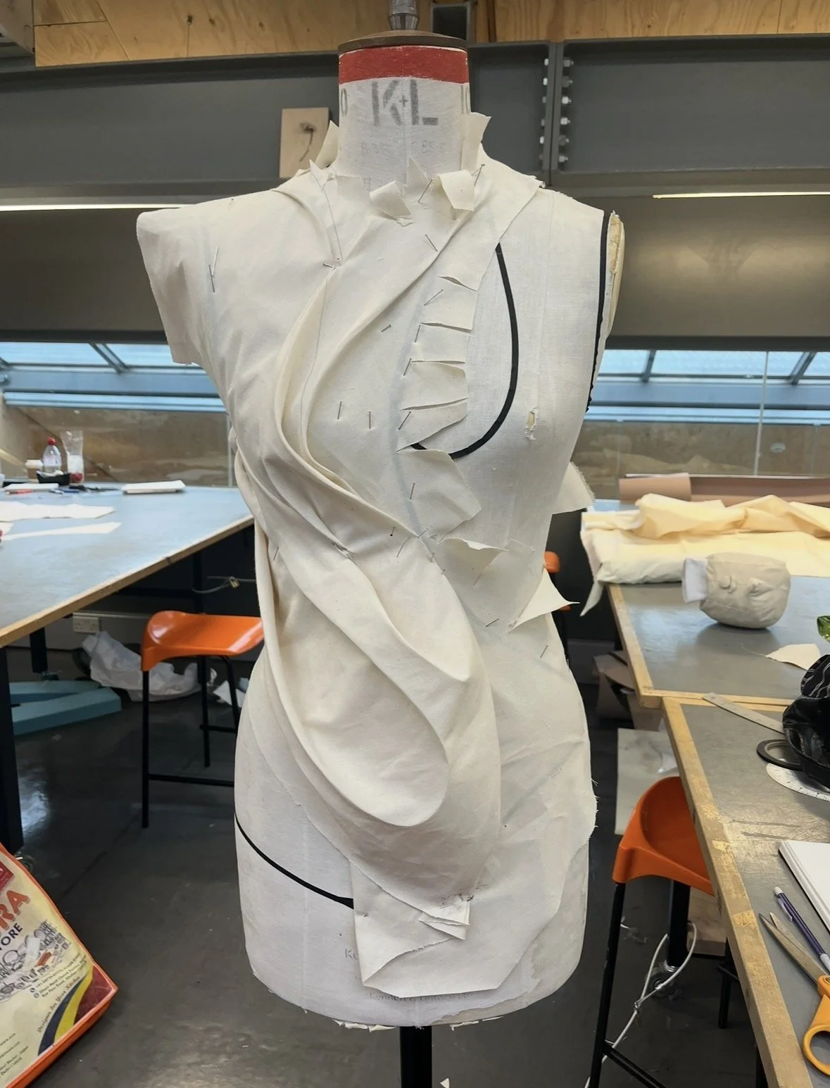 A dress form mannequin with a fabric pattern partially stitched, situated in a workspace with tables, stools, and supplies, used for fashion design or tailoring.