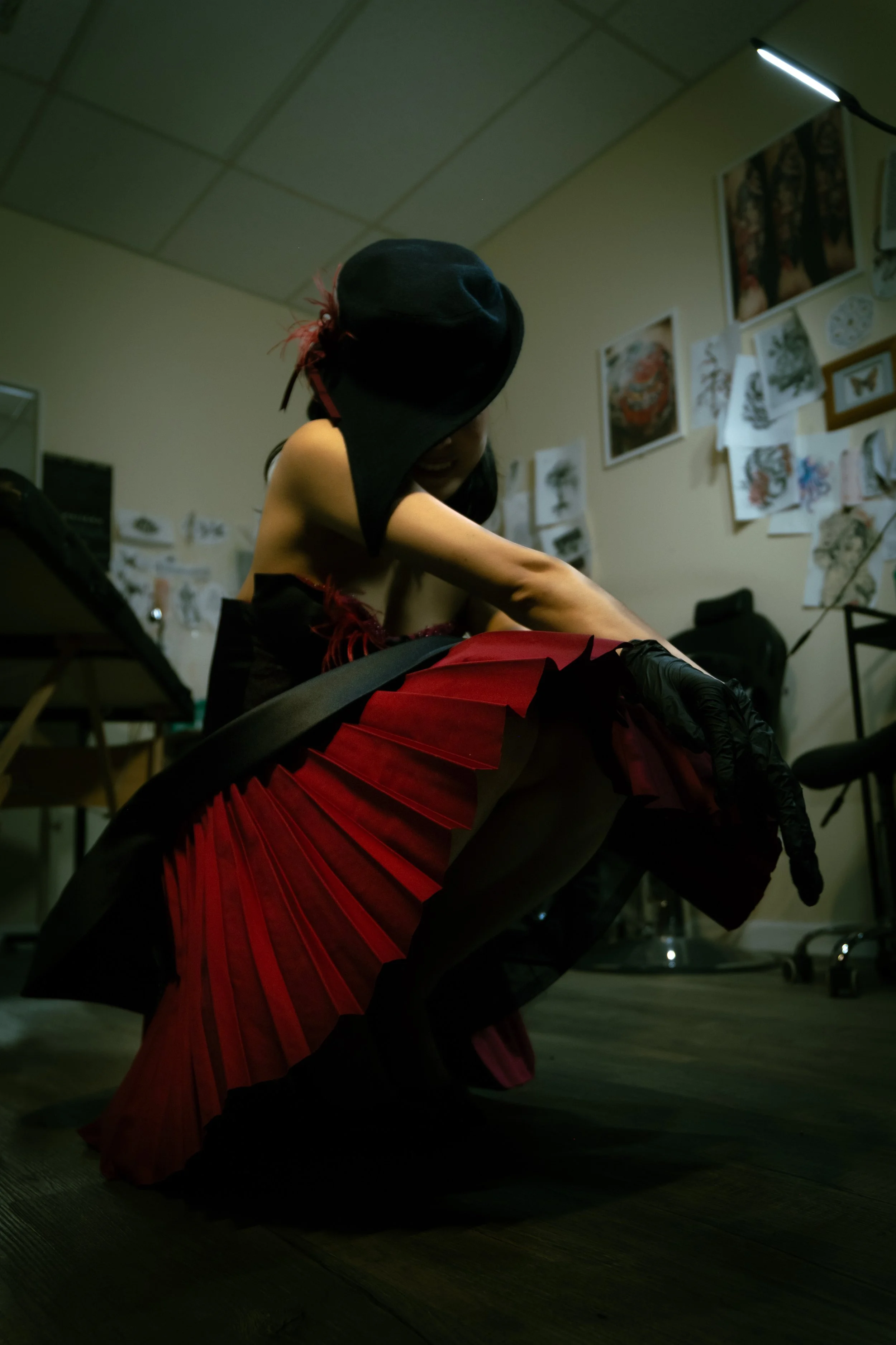 A person wearing a black wide-brimmed hat with red feathers, red and black attire with a pleated skirt, is bending over a table in an art studio. The background features various artworks and sketches on the walls, and the room is lit with a ceiling-mounted light.