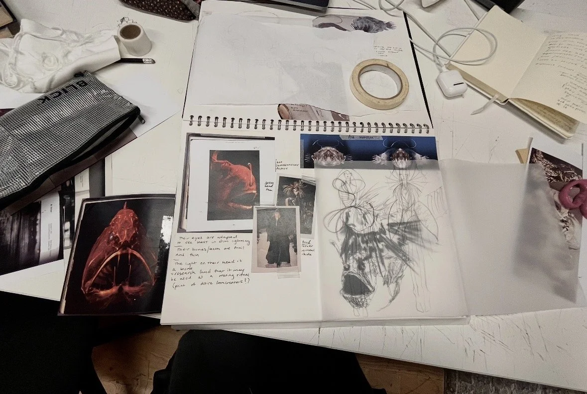 An overhead view of a cluttered white desk with sketches, photographs, a notebook, a roll of tape, a cup of coffee, a silver pencil case, and various cords and devices.