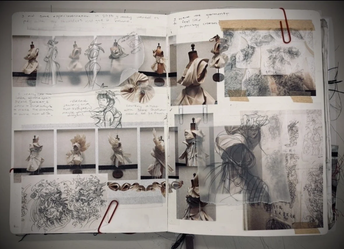 An art journal or design notebook containing sketches, fabric swatches, photographs of dress mannequins, and detailed costume designs, some annotated with handwritten notes and doodles. Red paper clips hold some pages together.